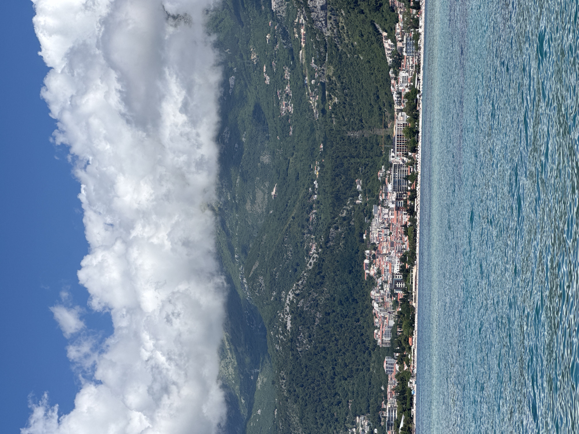 The image presents a panoramic view of Budva, Montenegro, a coastal city nestled between a large, verdant mountain range and the clear blue Adriatic Sea.
In the upper-left, a bright blue sky is visible, partially filled with large, white, billowy clouds. These clouds extend across the mountain peaks, casting some areas of the higher slopes in shadow or a misty embrace.
The central and left sections of the image are dominated by a steep, densely forested mountain. Its slopes are covered in lush green trees, with numerous buildings, likely houses and smaller settlements, interspersed throughout the lower and mid-sections, demonstrating human habitation extending up the natural terrain.
At the base of the mountain, along the narrow coastal strip, lies the city of Budva. A dense array of buildings, including residential structures and multi-story complexes, stretches along the coastline. Many buildings exhibit light-colored facades and traditional red-tiled roofs.
To the right, the calm expanse of the Adriatic Sea stretches into the distance. The water displays varying shades of blue and green, appearing clear with gentle ripples on its surface, indicating a tranquil body of water.
The scene captures a bright daytime setting, likely mid-morning or early afternoon, under partly cloudy skies over the mountains but with clear conditions over the sea. No specific human activities or individuals are discernible due to the distant perspective. There is no visible text in the image.