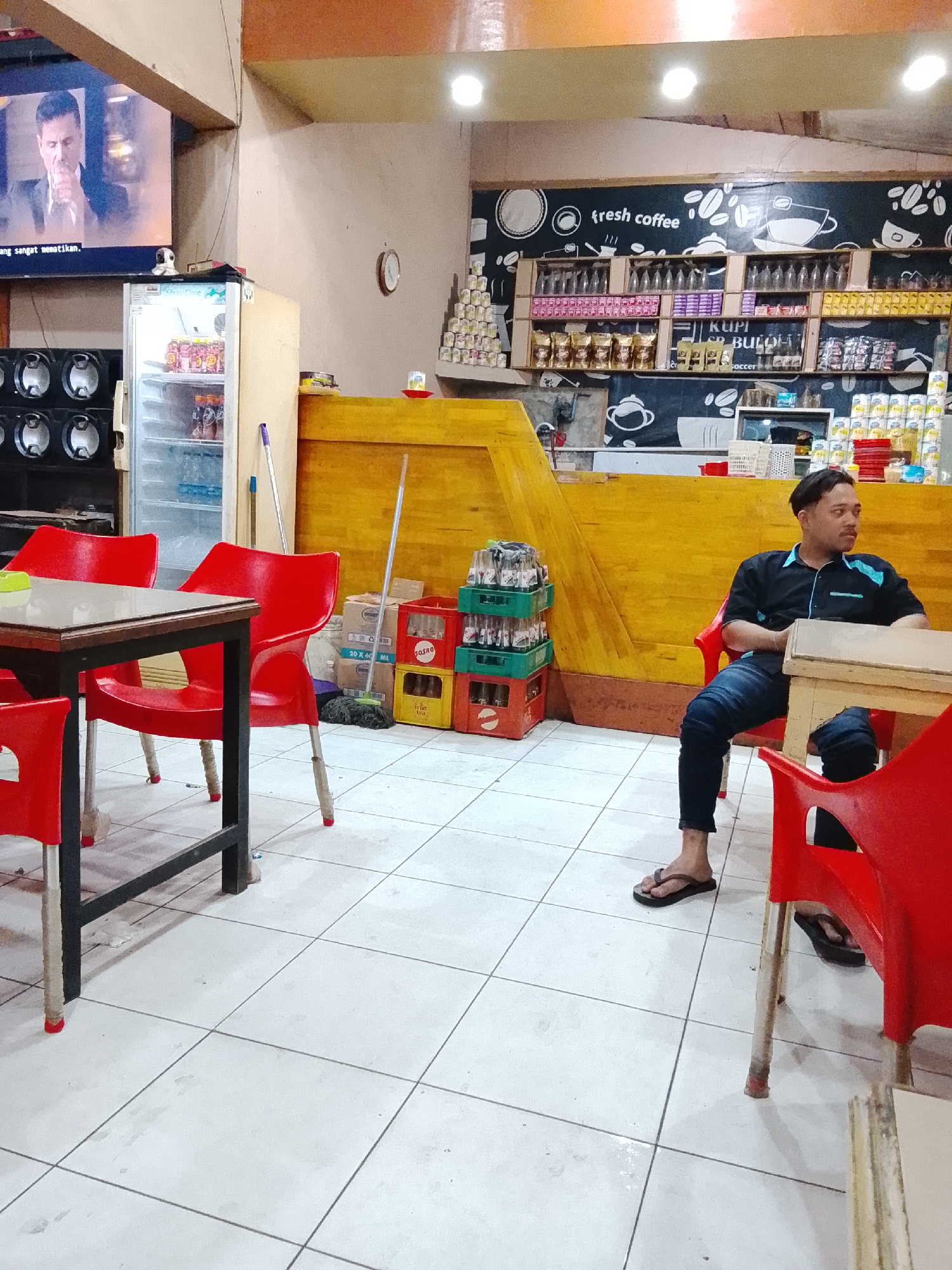 This image depicts the interior of a casual cafe or eatery in Lhokseumawe, Indonesia. The space features a light-colored tiled floor, several wooden tables, and bright red plastic chairs.
In the right foreground, a man with dark hair, wearing a black and light blue polo shirt and dark jeans, is seated at a table. He wears dark flip-flops and looks contemplatively towards the right, appearing to be resting or waiting.
Along the back and right walls, a yellow-paneled counter stretches, behind which a chalkboard-style wall mural displays white illustrations of coffee cups and beans, with the text "fresh coffee." Shelves are filled with various products, including bottled beverages, packaged goods, and cans. Some items display text such as "KUPI BUOL" (likely a coffee brand) and "Soccer." Cardboard boxes, one labeled "SOSRO" and "20 X 60 ML," and plastic crates of bottles are stacked on the floor, alongside a broom and mop.
On the left side of the room, a television mounted high on the wall shows a man in a suit with a serious expression, accompanied by a partial Indonesian caption reading "ang sangat mematikan." Below the TV, a white refrigerator stocked with drinks is visible, with a small security camera positioned on top. A large speaker system is situated next to the refrigerator. The cafe is illuminated by overhead lights, suggesting it's operating during the evening or night. The overall atmosphere is quiet and functional.