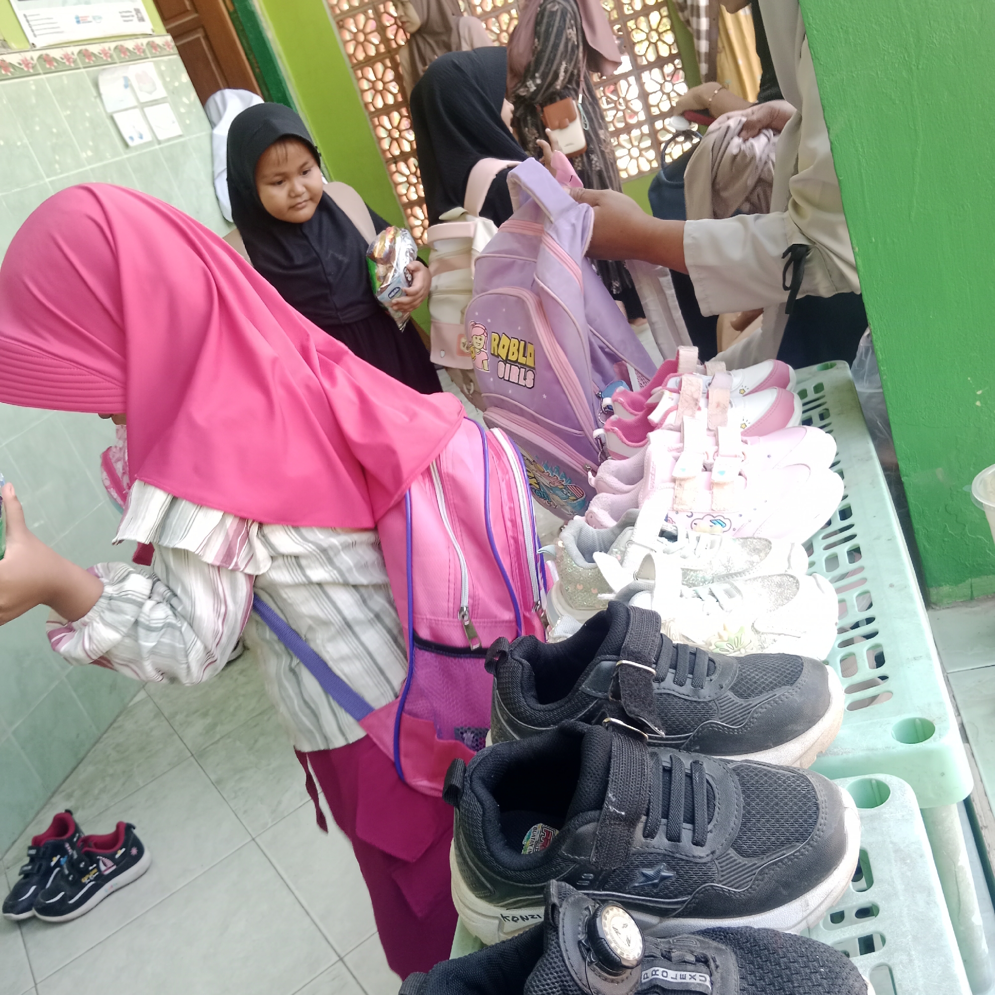 In Tegowanu Kulon, Indonesia, at a street-side stall, children and adults are engaged in a transaction. The primary focus is on children's footwear and backpacks laid out on a green plastic crate. Several pairs of sneakers, in various colors including black, white, and pink, are displayed for sale. Two purple backpacks, one with the "Roblox Girls" logo, are also visible. A young girl wearing a black hijab is holding a snack packet, while another individual, partially obscured by a pink hijab, is examining the merchandise. The background suggests an outdoor setting, possibly near a building with a green wall and decorative lattice work. The scene depicts a typical market exchange in a community.