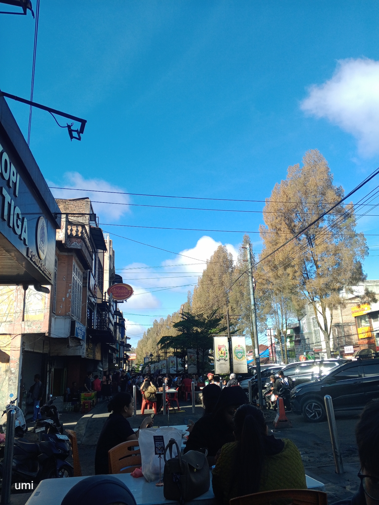 The image depicts a street scene in Berastagi, Indonesia, under a bright blue sky with scattered clouds. The foreground features a table with people seated around it, some holding phones. Beyond the table, a street is lined with buildings, many appearing to be shops and cafes. Signs visible include "KOPI TIGA" and "Golden Bakery & Cake Shop." Several vehicles, including motorcycles and cars, are present on the street, with some parked and others in motion. People are seen walking, sitting at outdoor tables, and interacting. Tall, bare-branched trees line one side of the street, their foliage appearing sparse. Electrical wires crisscross the sky above. The overall impression is of a bustling daytime urban environment.