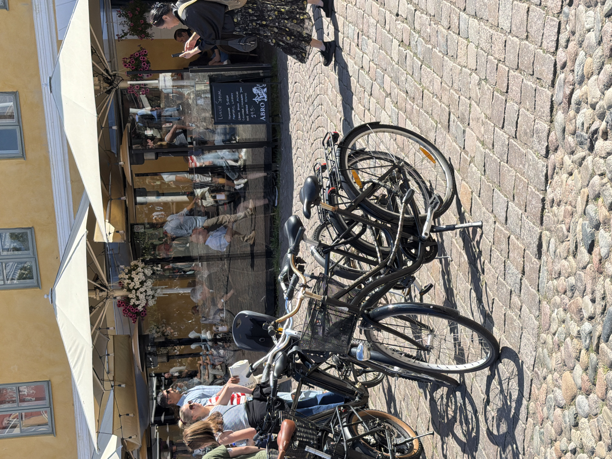 In Malmö, Sweden, on a sunny day, a cobblestone street is lined with parked bicycles. Beyond the bicycles, a yellow building forms the backdrop, with large windows reflecting the surrounding scene. Underneath awnings, an outdoor seating area of a restaurant is visible, with patrons dining inside. A woman, wearing a dark floral dress and carrying a bag, is captured in the process of taking a photograph with her smartphone. The scene suggests a lively urban environment with a focus on outdoor dining and cycling culture.