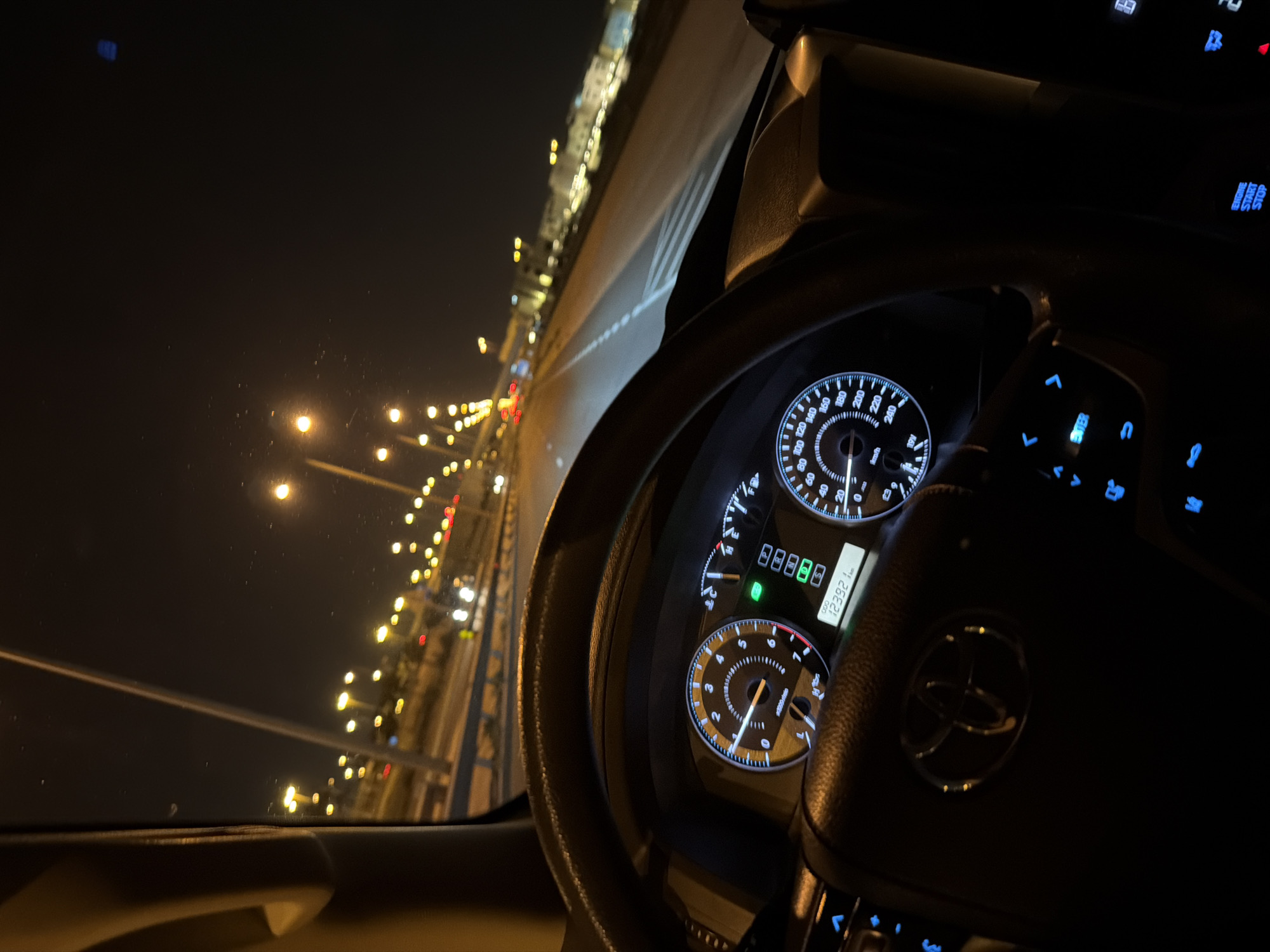 The image captures a night-time view from the driver's seat of a Toyota vehicle, as indicated by the prominent Toyota logo on the steering wheel. The dashboard is illuminated, showing a tachometer, speedometer, and a digital display indicating an odometer reading of 123921 km. The steering wheel controls are visible, some with blue illuminated buttons. Outside the car, the road stretches ahead, lined with streetlights and buildings, suggesting an urban environment. The overall scene depicts a person driving at night in a city.