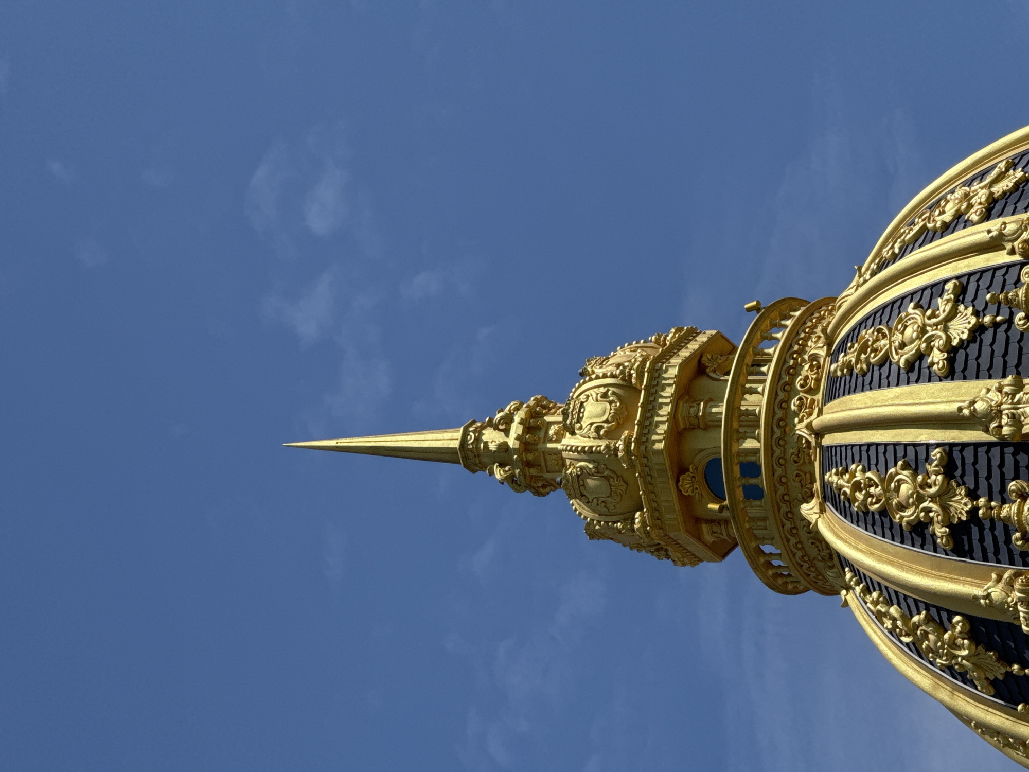 This is a close-up, low-angle shot of the golden spire and dome of a building, set against a clear blue sky with faint white clouds. The dome is covered in dark grey shingles and ornate golden decorative elements. The spire is tall and pointed, also made of gold, and is topped with a smaller finial. The image does not provide any information about people, actions, or interactions. The location is not identifiable from the image itself as Da Lat, Vietnam.
