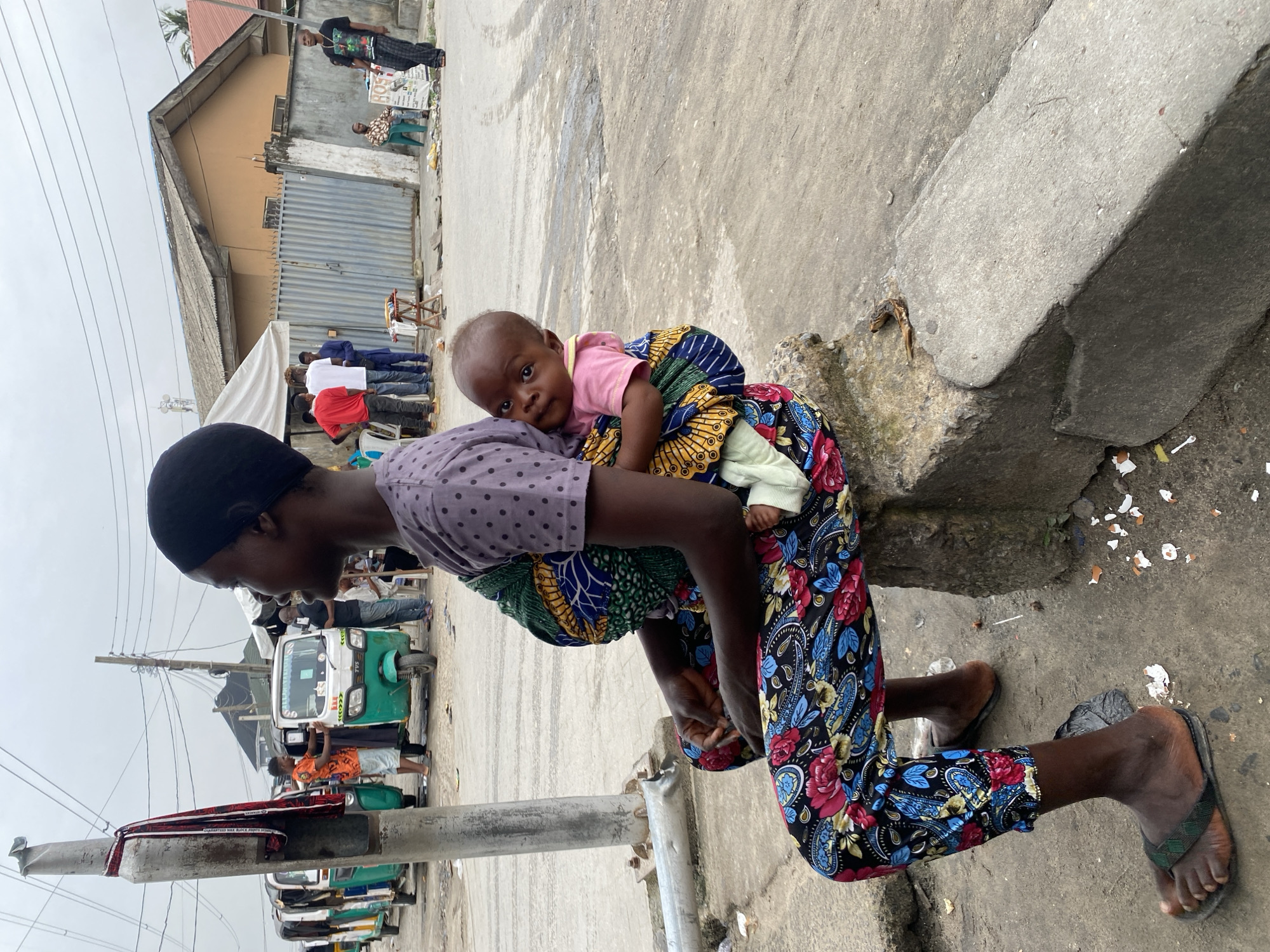 A photograph taken in Ado, Nigeria, depicts an African woman kneeling on a concrete sidewalk, carrying a baby secured to her back with a colorful fabric wrap. The woman wears a purple polka-dot shirt, printed pants featuring a floral pattern of red and blue on a black background, a dark head covering, and dark flip-flops. The baby wears a pink shirt and has one visible hand covered in a light-colored fabric. The woman is facing away from the camera but angled towards the viewer, with the baby looking directly at the camera. The background includes a building with a metal gate, people standing in a group, and several auto-rickshaws (Tuk-Tuks) parked along the street. Trash and debris are scattered on the ground. A utility pole is visible in the foreground, partially obstructing the view of the street scene. The sky appears overcast.