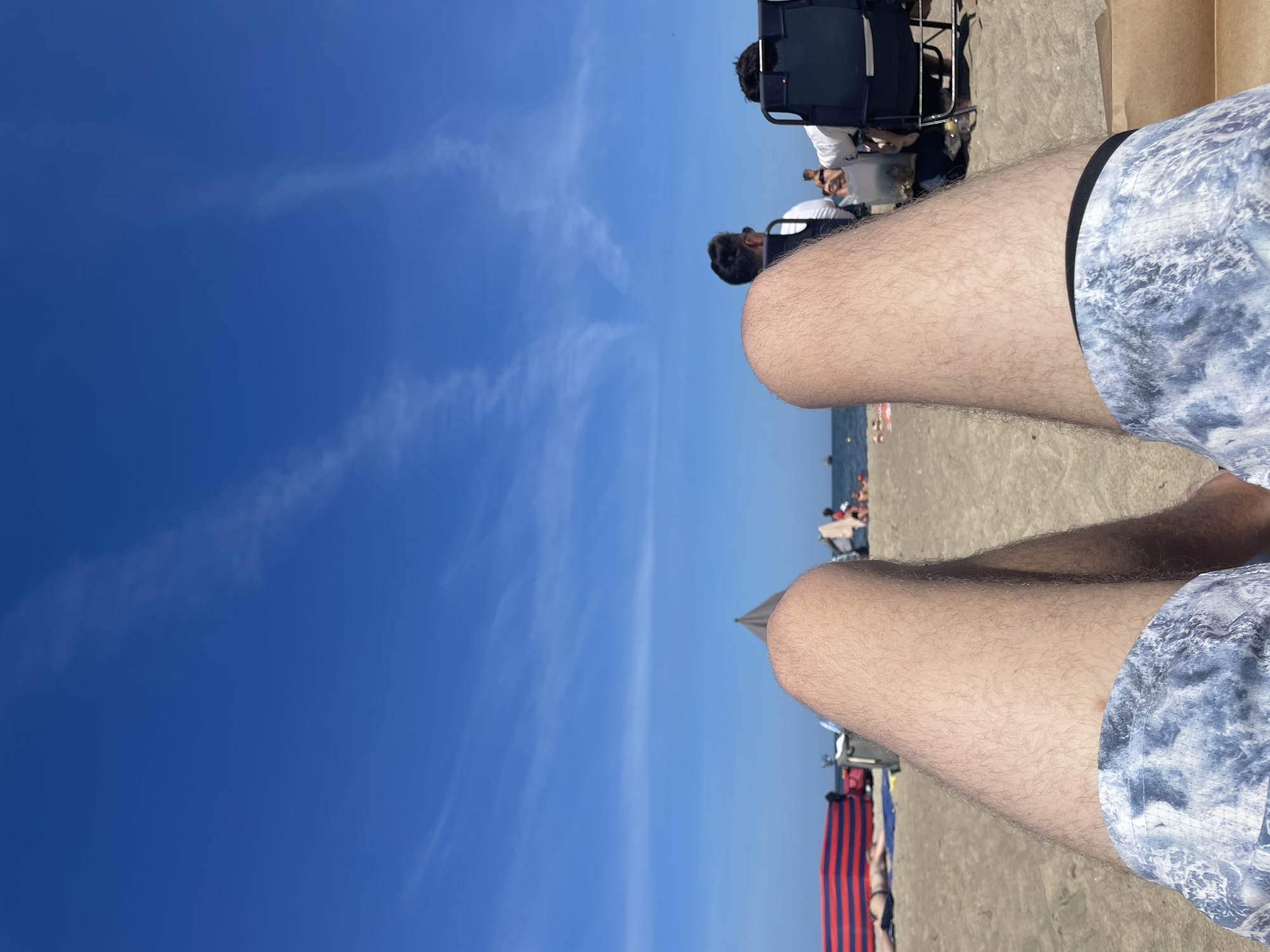 A first-person perspective shot shows a person's legs and shorts on a sandy beach. The sky is blue with some wispy white clouds. In the background, there are several beach chairs, umbrellas, and people. The beach is located in Sopot, Poland. The image captures a sunny day with a relaxed atmosphere.