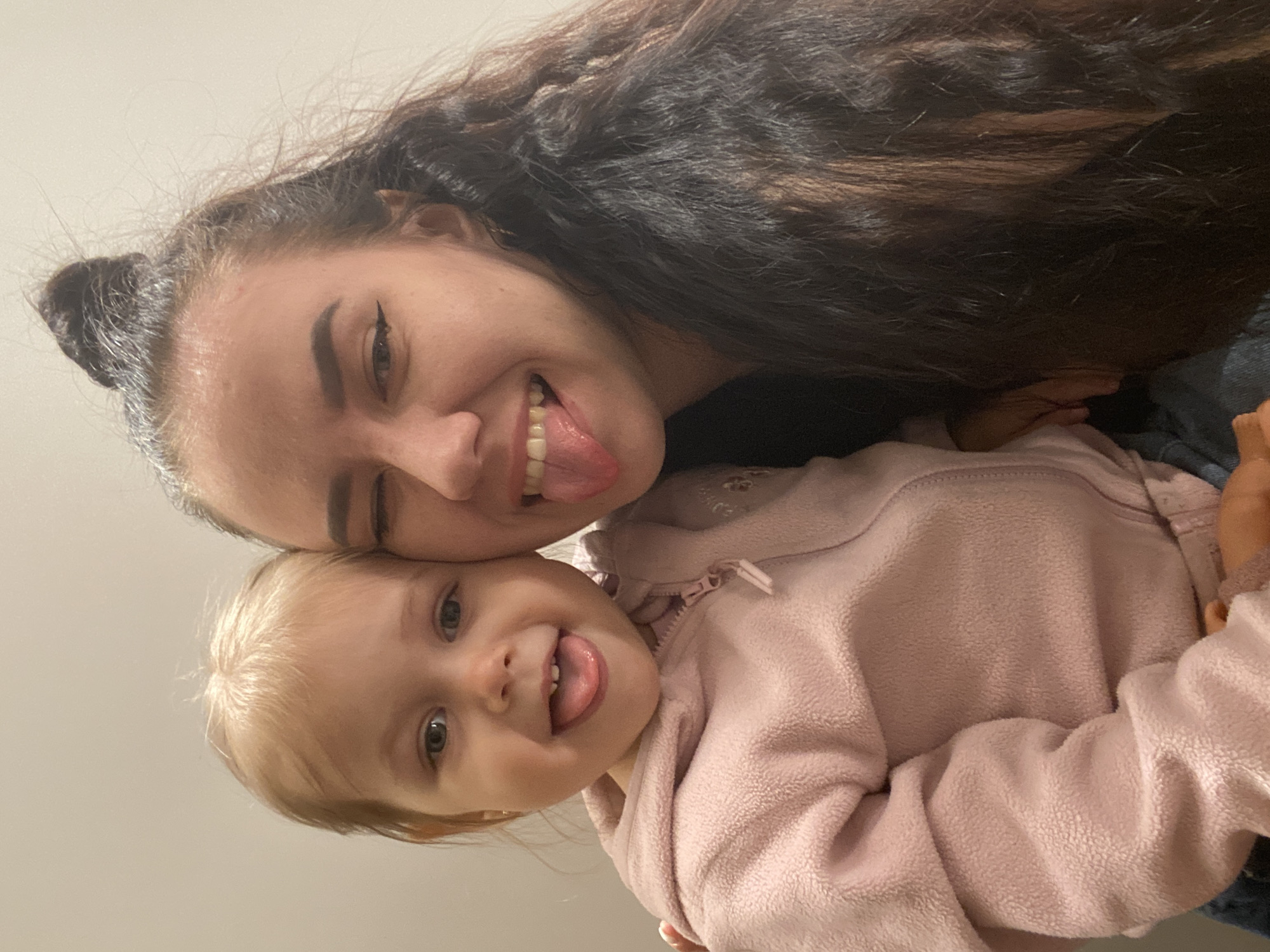 Two individuals are present in the image: an adult female and a young child, likely a toddler. Both are sticking out their tongues and smiling, indicating a playful or humorous interaction. The adult female has dark hair, styled with some of it pulled up into a bun on top of her head. She has noticeable eyeliner and fair skin. The child has blond hair and is wearing a pink fleece jacket with a zipper. The setting appears to be indoors, based on the presence of a wall as a background element. There are no indicators of the city of Perm, Russia.