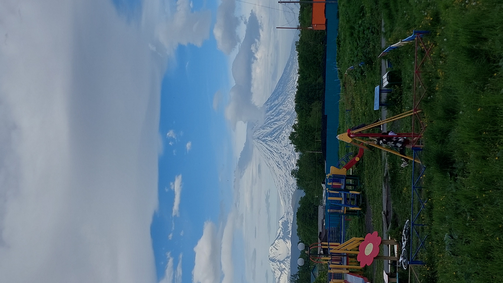 The image shows a playground in front of a snow-capped mountain. The sky is blue with some white clouds. The playground equipment is colorful, with a red and yellow swing set in the foreground and a blue and yellow climbing structure in the background. There is green grass and some yellow flowers around the playground. The mountain in the background is large and imposing, with snow covering its peak. The image appears to have been taken on a clear day, possibly in the late afternoon or early evening, given the lighting. The overall mood of the image is peaceful and serene, with the natural beauty of the mountain contrasting with the playful atmosphere of the playground. The location is likely Зелёный, Russia, as indicated by the context provided. There is no visible text in the image.