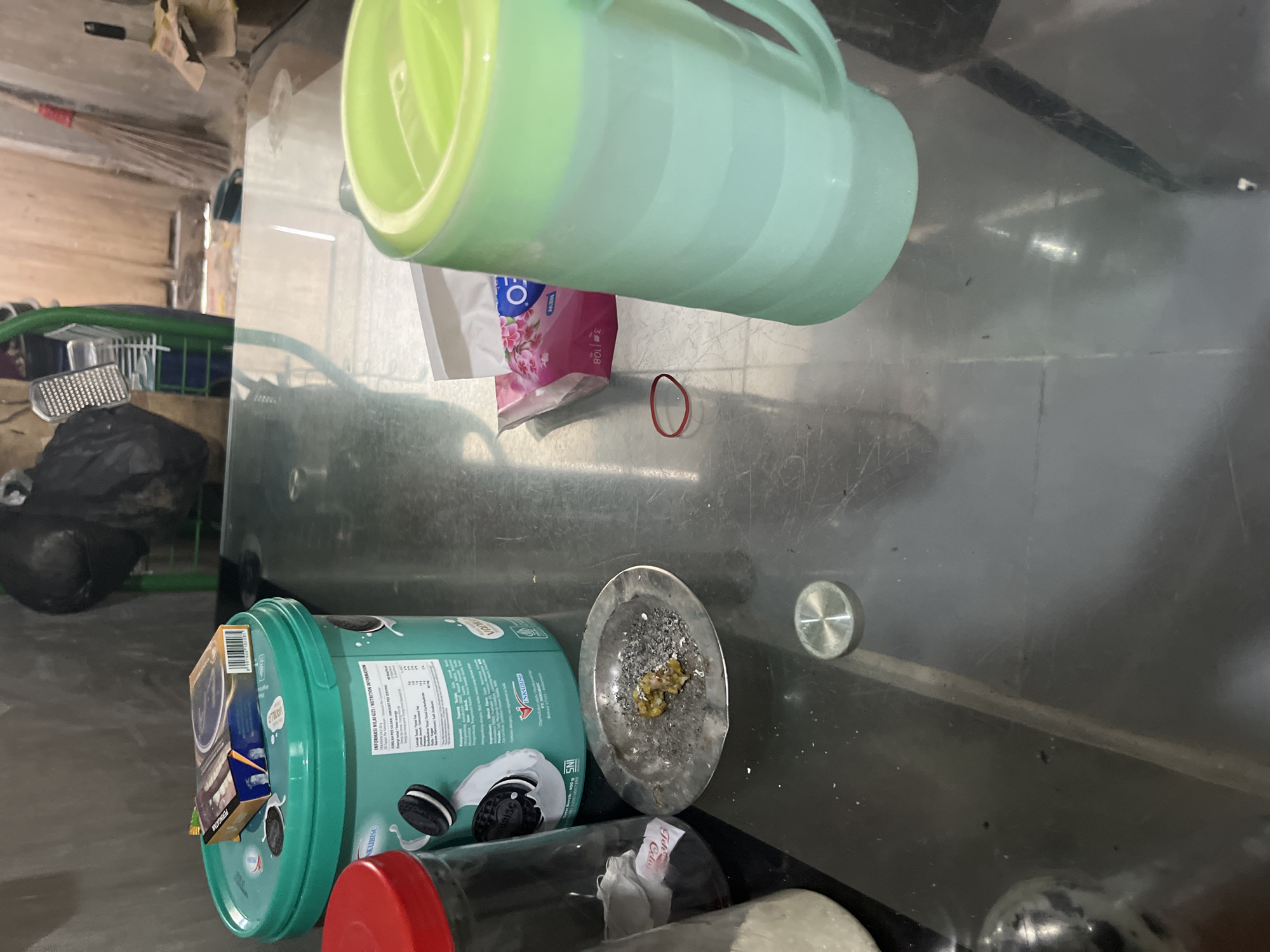 The image depicts a close-up, eye-level view of a dark, reflective surface, likely a table, in an indoor setting. The background is blurred, showing a concrete wall and what appears to be a broom leaning against it.
In the foreground and mid-ground, several objects are arranged on the table. To the left, a large teal bucket with an Oreo logo is prominent. A red-lidded glass jar with white contents is partially visible next to it. On top of the bucket rests a cigarette pack.
Towards the center of the image, a small, circular metal ashtray contains cigarette ashes and what appears to be a small, discarded piece of food or plant matter. A red rubber band is lying on the table near the ashtray.
To the right, a tall, light green plastic pitcher with a handle and a green lid is positioned. Behind the pitcher, a package of pink facial wipes is visible.
The lighting is somewhat dim, with reflections from the surface obscuring some details. The overall impression is one of a casual, domestic environment. No people are visible in the image.