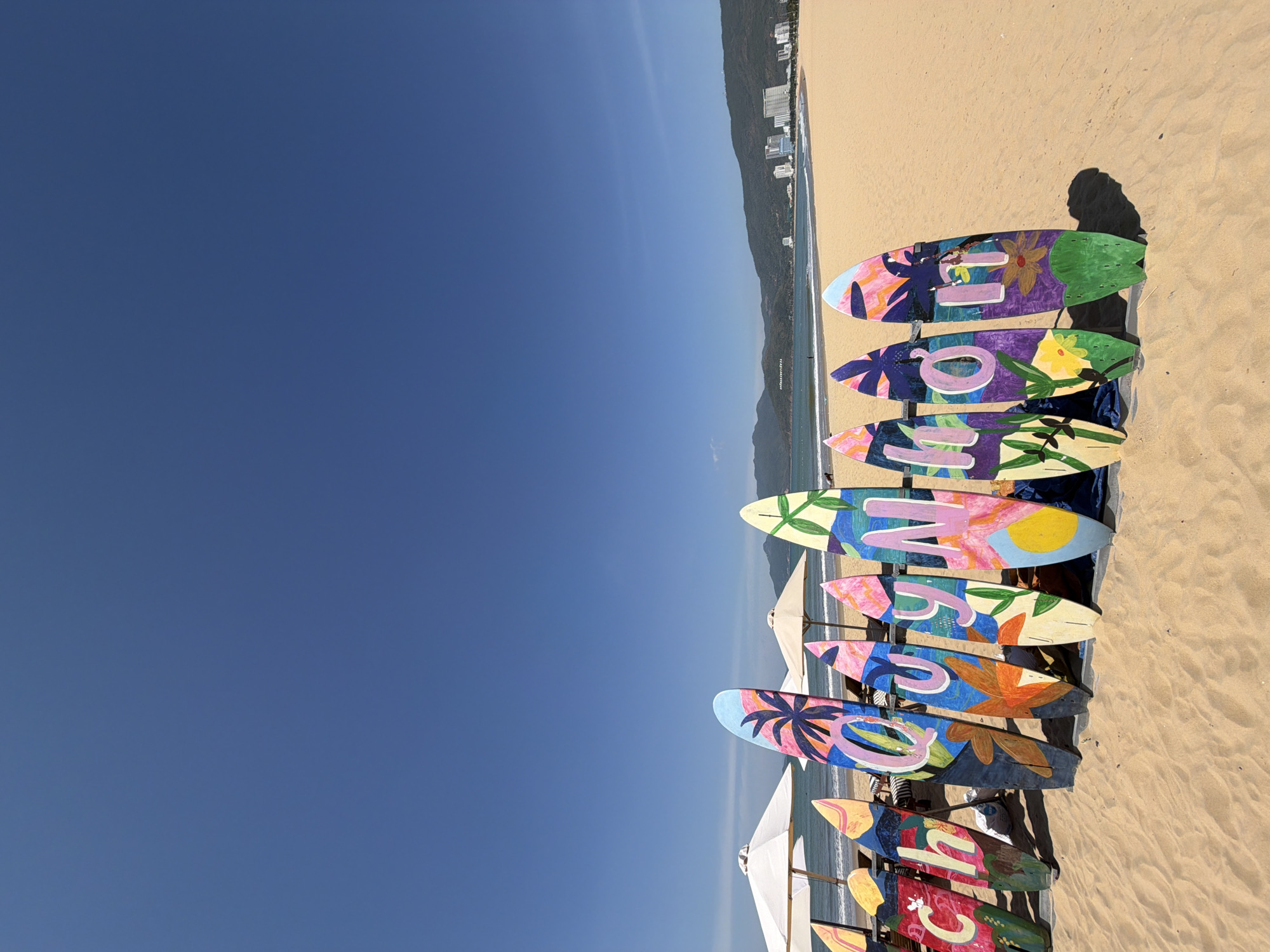 The image depicts a beach scene in Quy Nhon, Vietnam, under a clear blue sky. In the foreground, a row of colorful, hand-painted surfboards stands upright on the sandy beach. The surfboards are decorated with various patterns, including palm trees, flowers, and abstract designs, and some have lettering on them. Behind the surfboards, beach umbrellas are set up on lounge chairs, and the ocean stretches out to the horizon. In the distance, a coastline with green mountains and modern buildings is visible. The overall impression is a vibrant and sunny beach day.