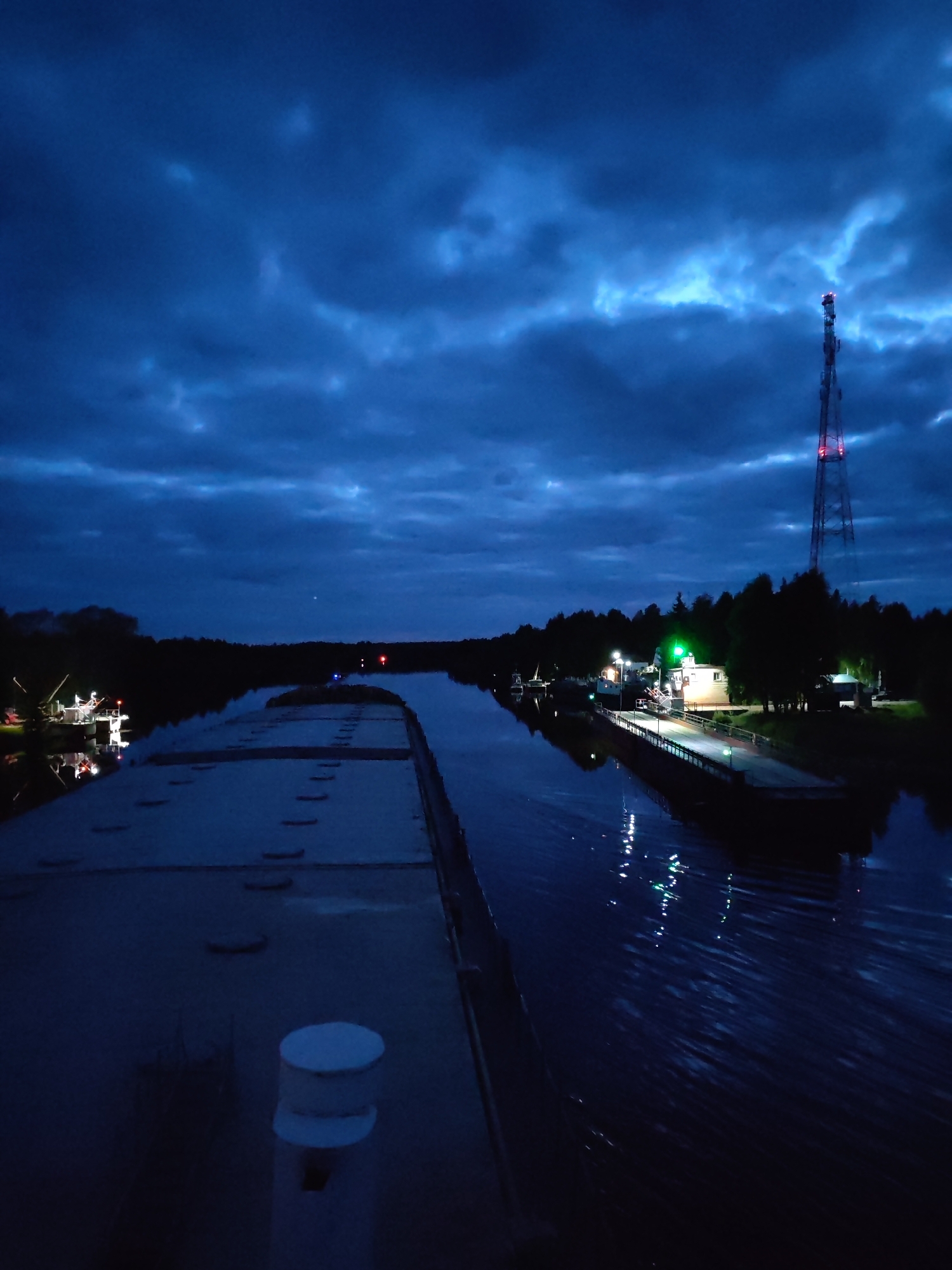 The image depicts a nighttime scene at Annensky Most, Russia. A canal flows through the scene, reflecting lights from surrounding structures. A barge occupies the foreground, its deck featuring hatches and a bollard. On the right, a small harbor is illuminated with lights. A communication tower stands in the background, its red and white pattern visible against the dark sky. The sky is filled with dark, layered clouds. The overall illumination is low.
