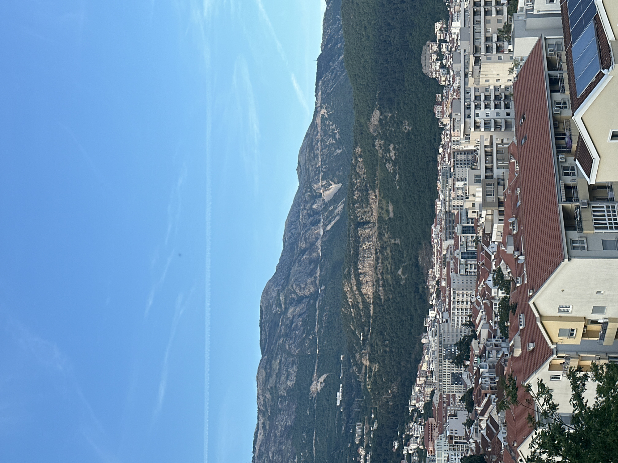 The image shows a view of Budva, Montenegro, with the imposing Lovćen mountain range in the background. The city's urban landscape features a mix of building styles and density, including modern apartments with red tiled roofs. The sky is clear blue, with contrails visible. The foreground offers a partial view of residential buildings.