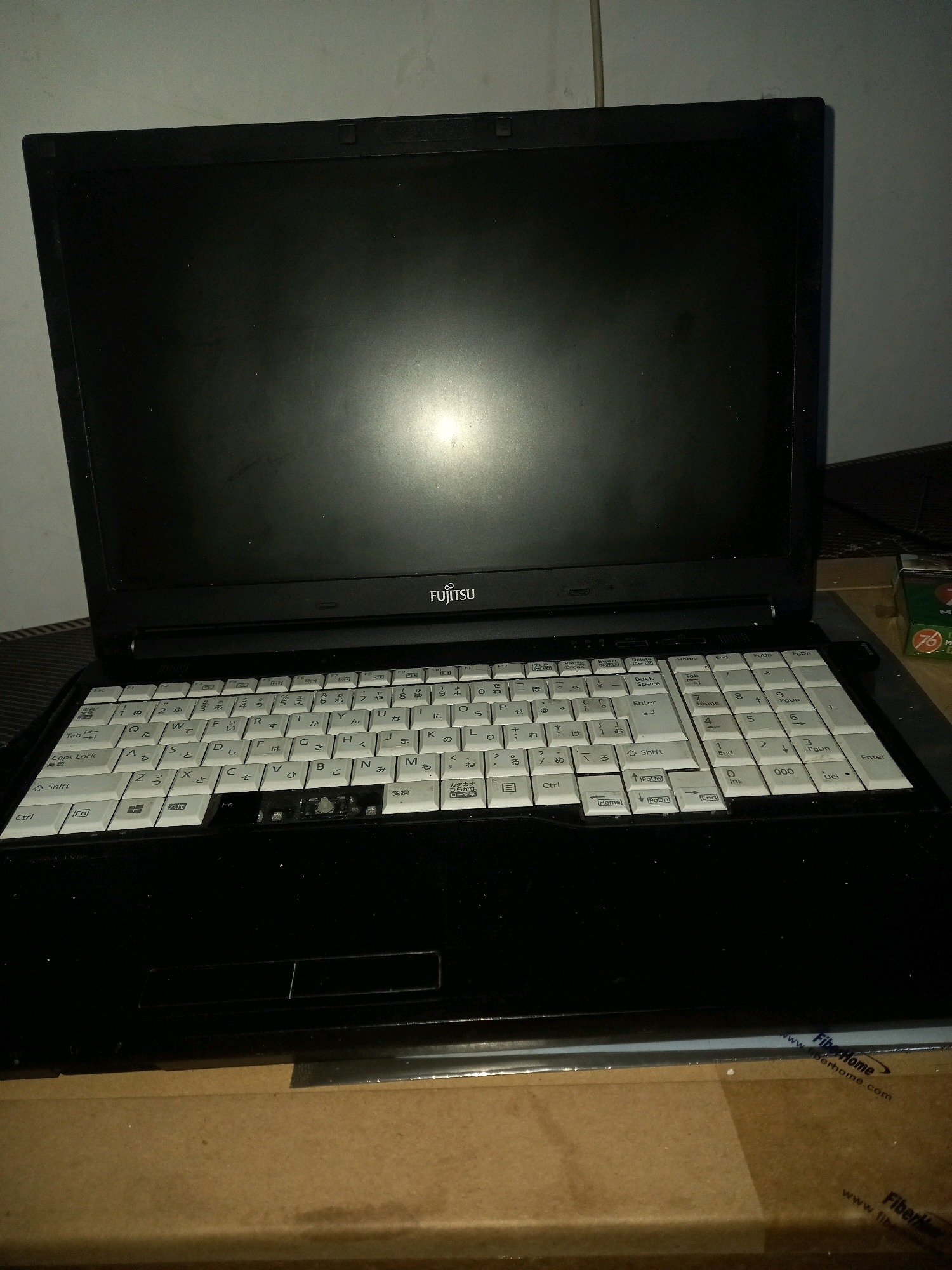 The image shows a black Fujitsu laptop computer, open and placed on a cardboard box. The laptop is the primary subject. The keyboard is visible and appears to be a Japanese layout with Hiragana and Katakana characters. The screen is dark, reflecting some ambient light. To the right of the laptop, a small green and white box with red lettering and the number "76" is partially visible. The background consists of a plain wall, partially shadowed. The overall environment appears to be indoors, likely a room. The image is taken from a slightly elevated angle looking down at the laptop. The location is described as Mejasem Timur, Indonesia, but there are no specific visual cues in the image to confirm this geographical location.