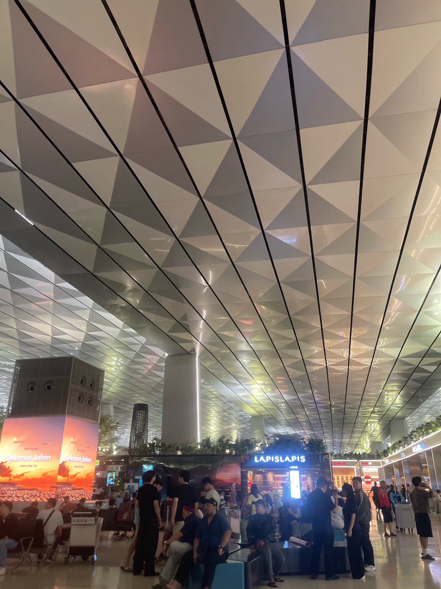 The image depicts an interior view of an airport terminal, likely Soekarno-Hatta International Airport in Tangerang, Indonesia, not Pajang. The ceiling is a prominent feature, characterized by a geometric pattern of triangular panels with alternating light and dark shades, separated by thin black lines. The lighting in the terminal is a mix of ambient light reflecting off the ceiling and illuminated signage and screens.
In the foreground and midground, there are several groups of people in a waiting area. They are dressed in casual attire, with some seated on benches and others standing. Luggage carts are also visible.
To the left, a large, illuminated digital display shows the phrase "Selamat Jalan" (meaning "Have a nice trip" in Indonesian) set against a sunset-like backdrop. Further into the terminal, a retail establishment with the prominent sign "LAPISLAIS" is visible, along with other shops and signs that are partially obscured. Greenery, in the form of plants, is integrated into the architecture, particularly along the upper edges of the structures and in vertical planters. The overall impression is a modern and well-lit airport environment.