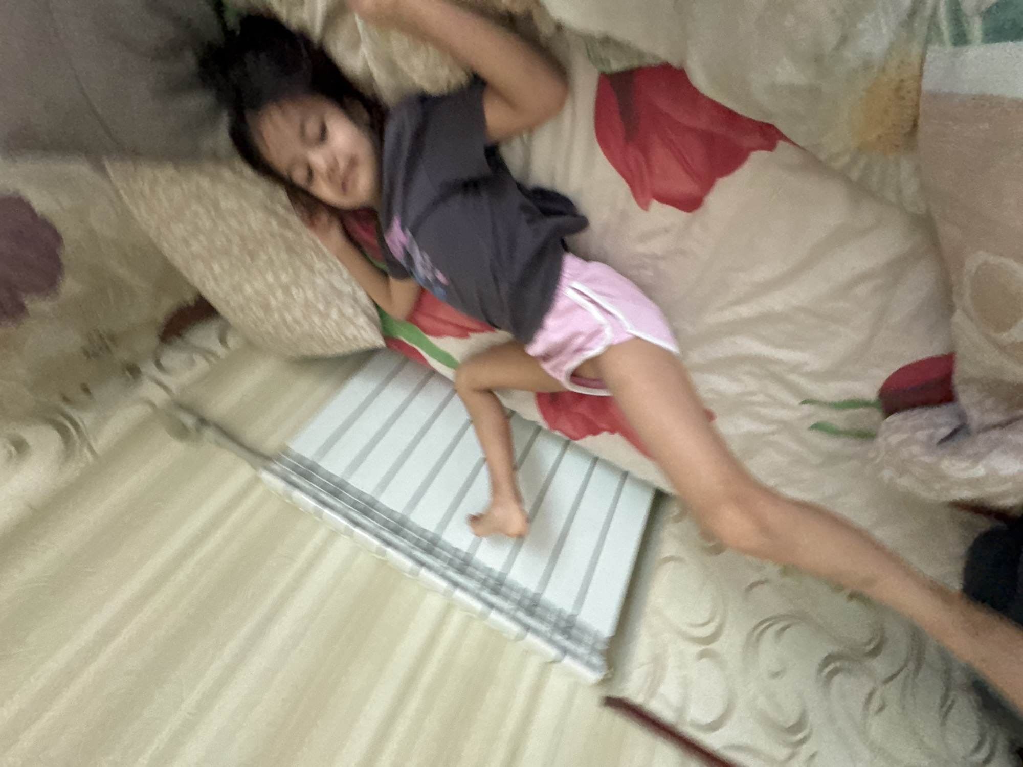 The image shows a young girl lying on a bed, seemingly asleep. She is wearing a dark grey t-shirt and pink shorts with white trim. Her head is resting on a pillow with a floral pattern, and she is covered by a blanket that also features a red floral design. The setting appears to be indoors, possibly a bedroom, with other soft furnishings visible. A striped object, which could be a mat or part of the bedding, is positioned next to the girl. The lighting suggests it might be daytime, though the exact time is unclear. The overall mood is peaceful, hinting at a moment of rest. There is no visible text in the image. The location context provided is Poltava, Ukraine.