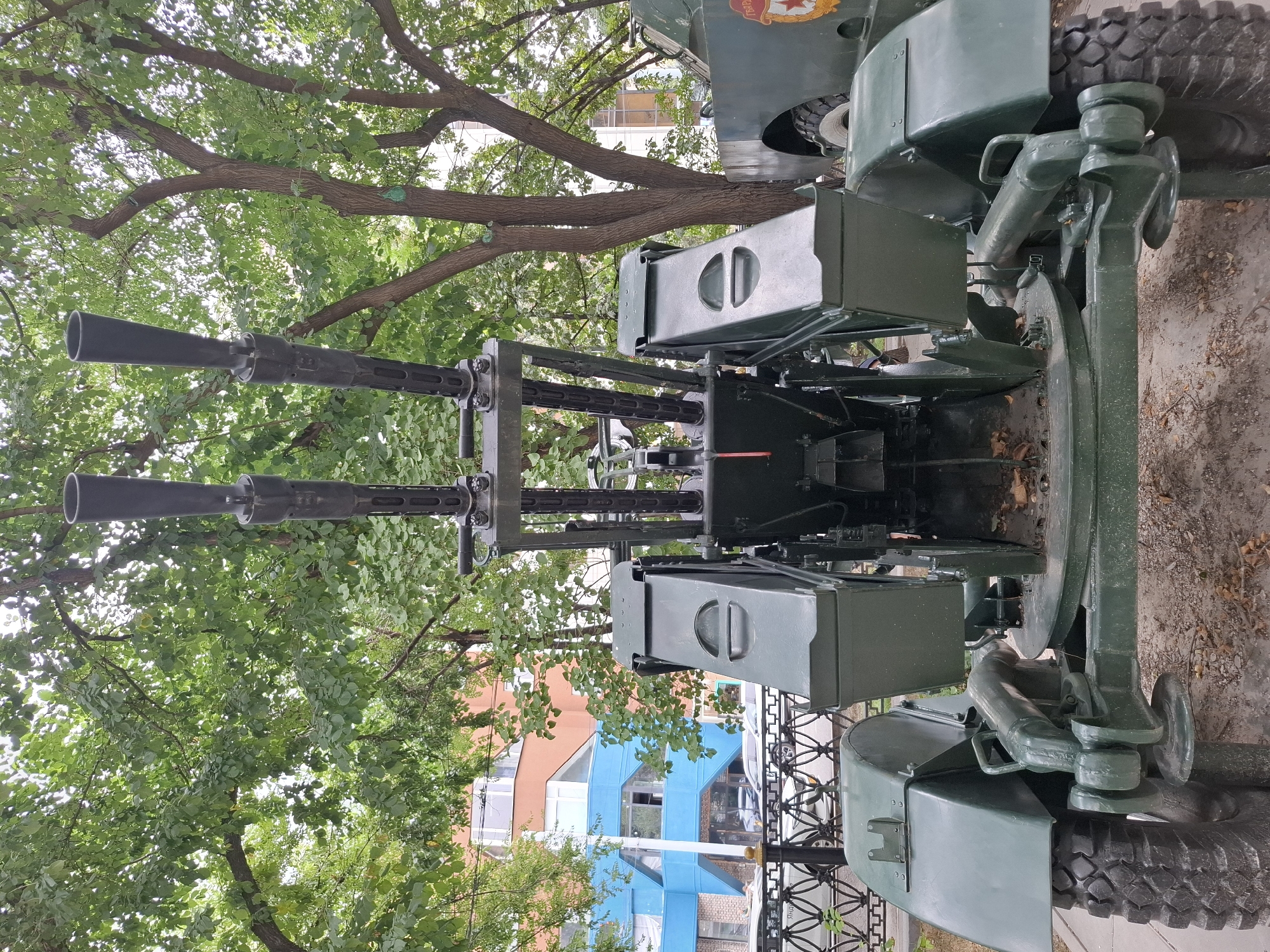 This is a full, eye-level, outdoor, medium shot of a twin-barreled anti-aircraft gun, painted in olive drab, on a wheeled mount. The gun barrels are pointed upwards and forward. The scene is framed by lush green trees with dappled sunlight filtering through the leaves. In the background, there is a building with colorful facades: one section is orange and the other is bright blue. A black metal fence runs along the bottom of the image, and beyond it, cars are visible. The ground appears to be a mix of dirt and fallen leaves. The overall atmosphere is one of a static display, likely in a public space within the city of Almaty, Kazakhstan.