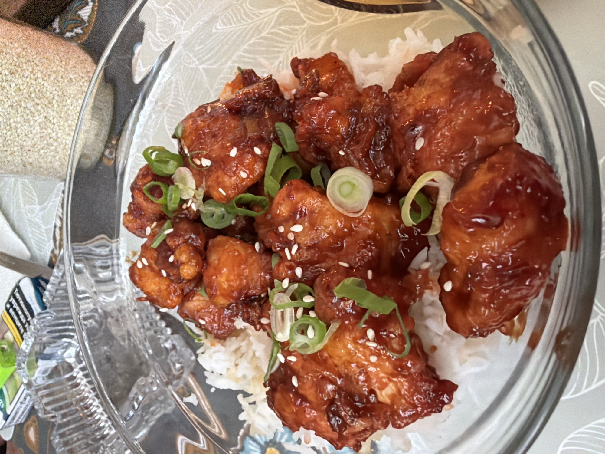 A bowl of cooked rice topped with glazed chicken and garnished with green onions and sesame seeds is presented in a glass bowl. A portion of the table setting surrounding the bowl is visible, with a patterned placemat underneath the bowl and part of a fabric item adjacent to the bowl. The setting does not offer sufficient information to determine the location within Sofia, Bulgaria.