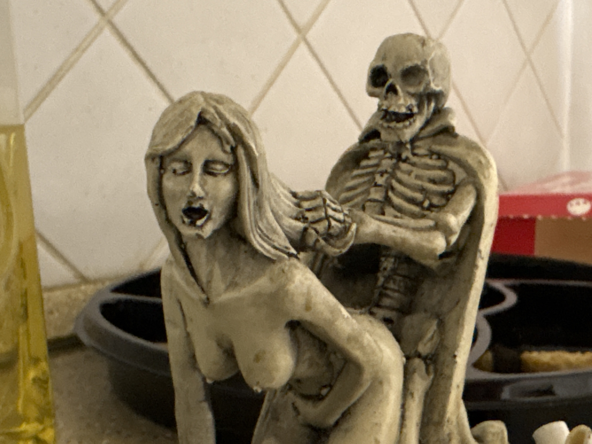 The image shows a figurine depicting a woman and a skeleton. The woman is in a bent-over position with her mouth open. The skeleton stands behind her with a hand on her shoulder. The background includes a kitchen setting with tile and other items on a counter. There is no evidence that the image was taken in Perm, Russia.