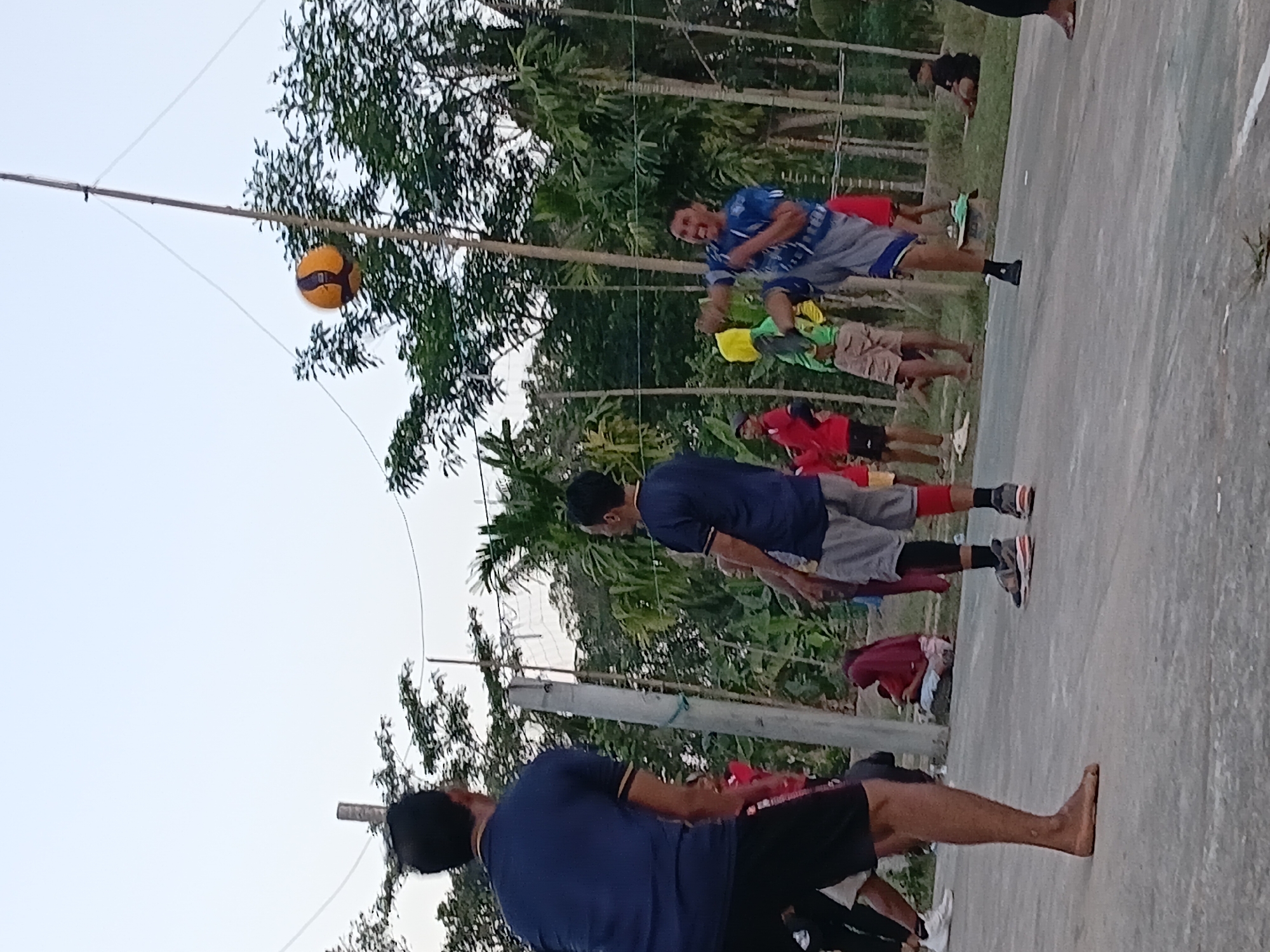 This is a vertical, medium-shot, outdoor photograph taken at dusk in Margo Rukun, Indonesia. The scene depicts a group of people engaged in a game of Sepak Takraw. In the foreground, a man on the left, seen from the back, is wearing a dark blue t-shirt and black shorts. He is barefoot and facing away from the camera, towards the action. To the right, a man in mid-air is performing a kick with his left leg, aiming for a volleyball suspended by a pole and wires. He is wearing a blue t-shirt with a graphic print and gray shorts with blue trim. His facial expression is one of exertion. Other individuals are visible in the background, some observing the game, others participating. The environment consists of an open, paved area, likely a court, surrounded by trees and vegetation, consistent with a rural or semi-rural setting in Indonesia. The sky is overcast and light.