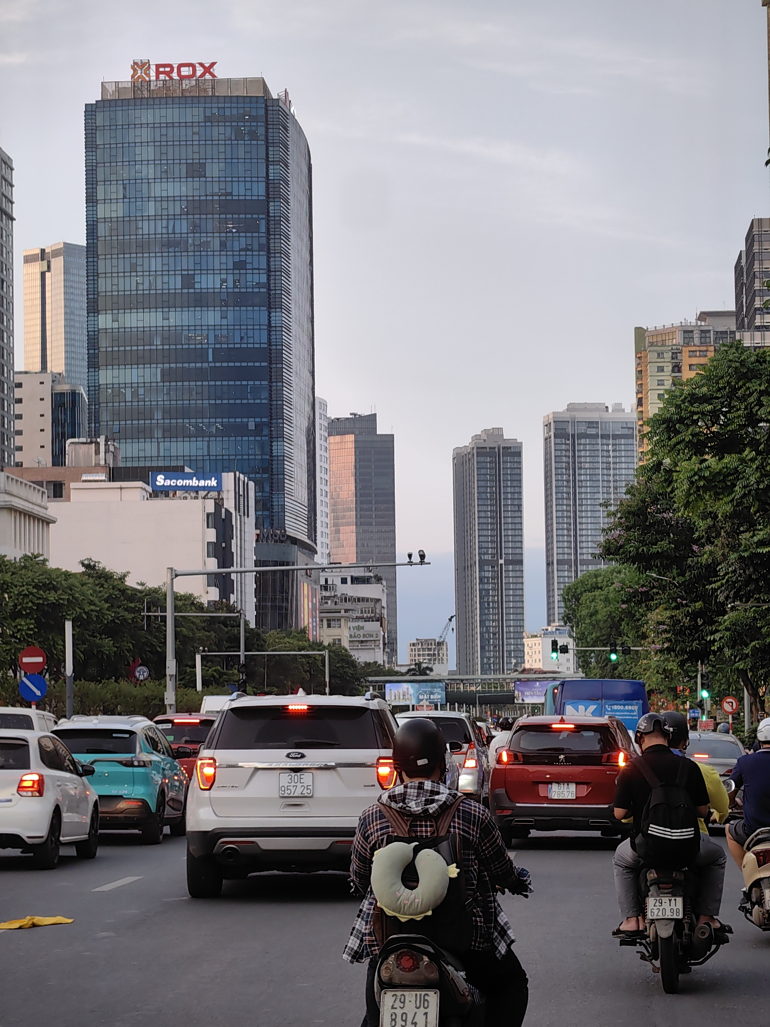 This eye-level shot captures a bustling city street in Hà Nội, Vietnam, under a soft, hazy sky, likely during late afternoon or early evening. The scene is dominated by a dense flow of traffic, consisting of numerous cars and motorbikes.
In the foreground, two motorbikes are prominent. The closest rider, wearing a plaid shirt and helmet, has a light green neck pillow attached to their backpack, and their motorbike displays license plate "29-U6 8941". To their right, another motorbike carries two individuals, both with helmets, showing license plate "29-V1 620.98". Cars fill the lanes, including a white Volkswagen Polo, a light blue SUV (possibly a VinFast VF e34), and a prominent white Ford Explorer SUV in the center with license plate "30E 957.25" and illuminated brake lights. Behind it, a red Peugeot SUV has the license plate "61A 785.76". Green traffic lights are visible further down the street, indicating ongoing movement.
The background features a modern urban skyline with several tall buildings. A prominent glass skyscraper on the left proudly displays a red "XROX" logo at its summit. Below it, on a smaller building, the "Sacombank" logo is visible, alongside a partial "MSB" sign. Other high-rise residential and commercial buildings extend into the distance. An elevated structure spanning the street displays various advertising texts such as "MẶT BIỂN", "TRÊN TANG", "4 VIỆN BẢO SƠN 2", and "1800.6800". The visible Vietnamese license plates and architectural style firmly place the scene in Hà Nội, capturing the dynamic atmosphere of a major Southeast Asian city.