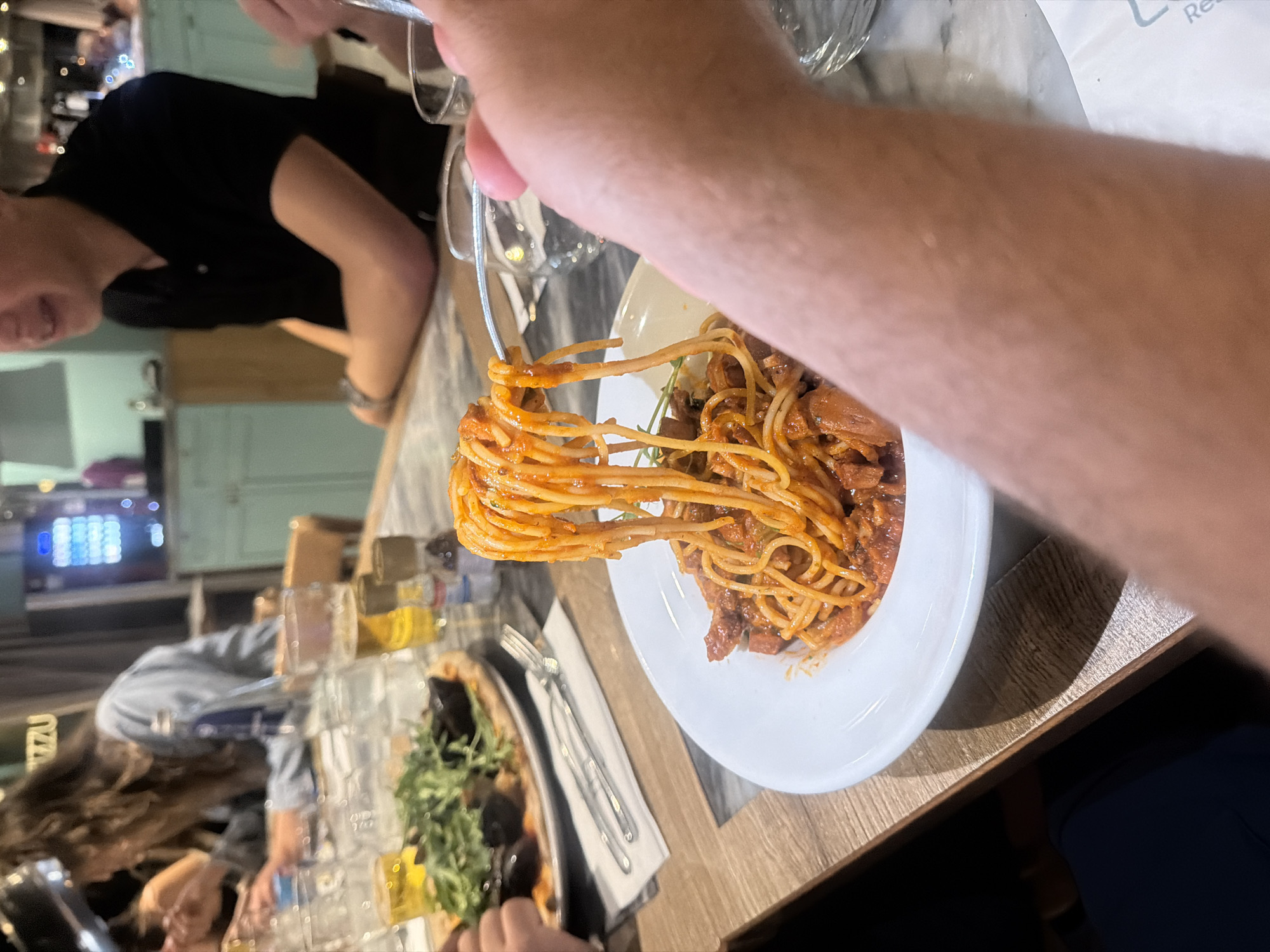 A person is twirling spaghetti onto a fork from a white plate of spaghetti with sauce. In the background, people are seated at a table, and there are glasses and food, including a pizza. The setting appears to be a restaurant, given the casual attire of the diners and the presence of multiple food items and tableware. The precise location in Saint Paul's Bay, Malta cannot be determined from the image alone.