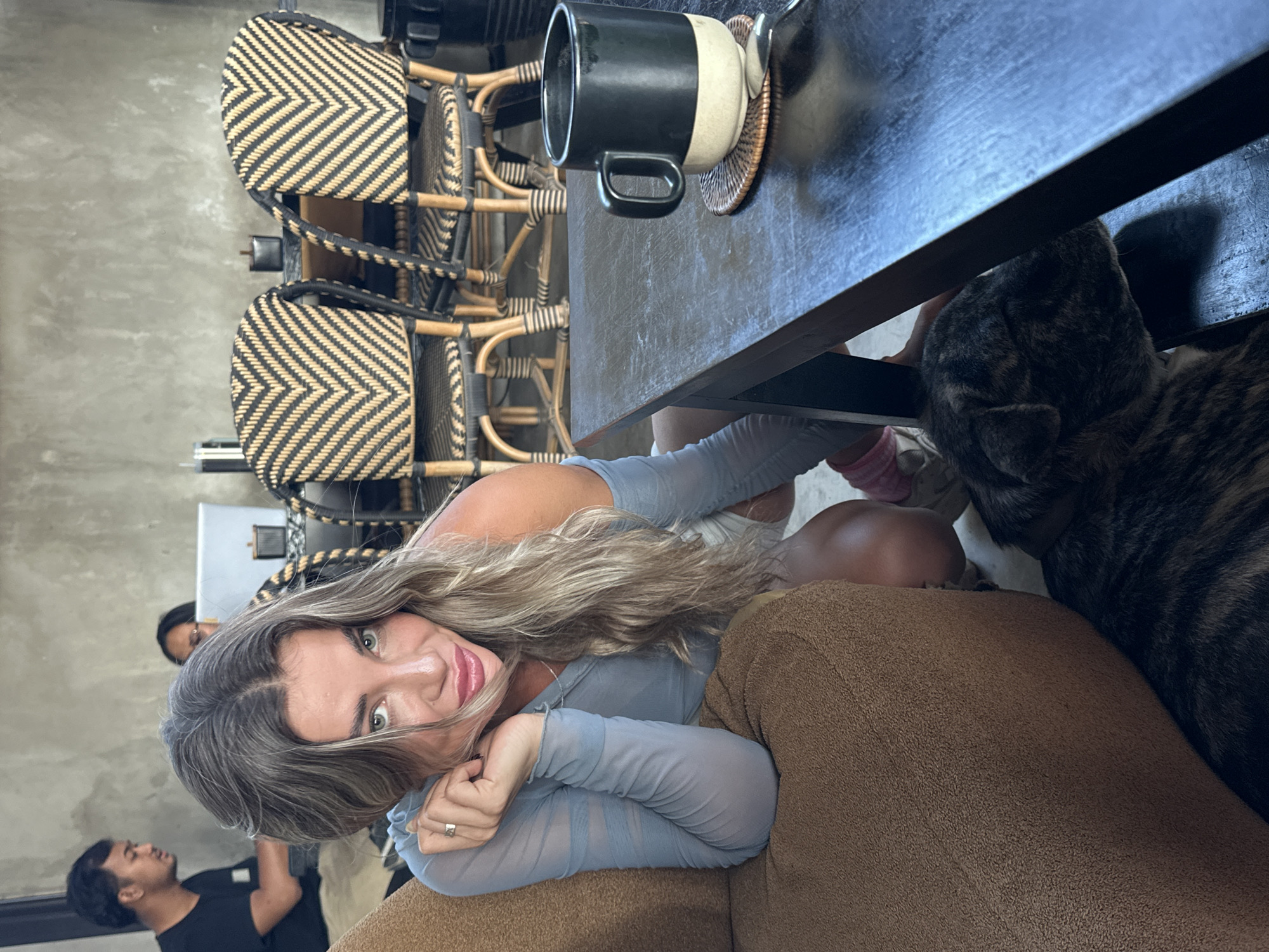 The image appears to be taken in a cafe or restaurant in Kuta Selatan, Indonesia, based on the decor, ambiance, and likely cultural context. A young woman with long blonde hair is prominently featured, resting on a brown upholstered couch or chair. She is wearing a light blue sheer top. Her arms are positioned with her hands near her face. The focus is on her face, which is angled towards the camera.
A dark brindle-colored dog is near her, partially visible under a dark-colored table. On the table, there is a large black and white mug on a woven coaster. In the background, there are several bistro-style chairs with a black and natural woven design, suggesting a casual dining atmosphere. Two men are further in the background, one wearing a black shirt and another partially visible. The lighting is soft and ambient, contributing to a relaxed and comfortable vibe.