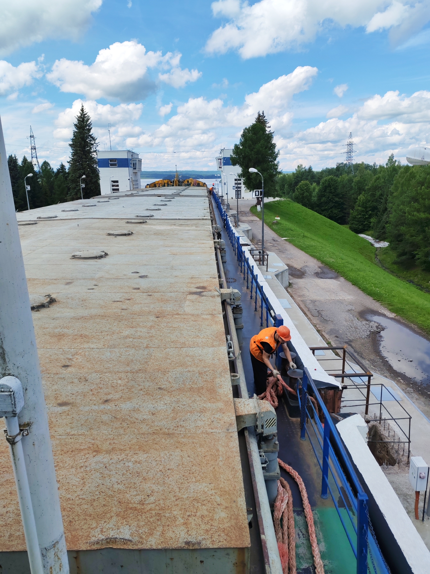 A person wearing an orange safety vest and helmet is securing a thick, pink rope to a bollard on the deck of a barge. The barge is docked alongside a concrete structure with a blue railing. In the background, there is a grassy slope leading up to a road and trees. Further in the distance, there are buildings with white and blue facades, as well as electrical pylons and a clear sky with white clouds. The scene appears to be a part of a dam or lock system, possibly in the vicinity of Белоусово, Russia, given the mention of a city.