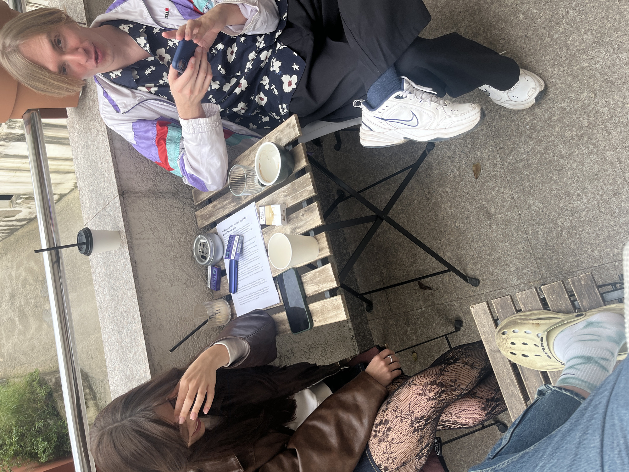 The image, captured from an oblique, slightly elevated perspective and rotated, depicts two individuals seated at a small wooden slatted table, likely on a balcony or cafe terrace in Budva, Montenegro. The flooring beneath is a light-colored speckled stone, and a metal railing is visible on the left side, suggesting an outdoor setting.
The person positioned towards the top-left of the frame has blonde hair and wears a multi-colored retro-style windbreaker over a navy shirt with white floral patterns and dark pants. They are wearing white Nike sneakers. This individual holds a small blue electronic device, possibly a heated tobacco device. Their expression is neutral, looking towards the viewer.
The second person, situated towards the bottom-right, has dark hair and wears a brown leather jacket. Their legs are visible, adorned with patterned black fishnet stockings. One foot, clad in a beige Croc shoe with a tie-dye patterned sock, rests on the wooden table. A simple ring is visible on their hand, which is held near their face, possibly resting or covering a smile.
The wooden table between them is covered with various items. These include a white paper cup with a black straw, a silver ashtray, two small purple and blue boxes labeled "TEREA," a blue electronic heating device (similar to the one held by the first person), a transparent glass, and a white ceramic mug containing some residue, possibly from coffee. Also on the table are a Marlboro cigarette pack, a smartphone displaying "6:23" (indicating time), and a white document with text, partially visible, which includes the phrase "1) Reasons Why Renting Could Be Better Than Buying." The overall lighting suggests a mild or overcast day, contributing to a relaxed atmosphere. No strong emotions are overtly displayed, implying a casual social gathering.