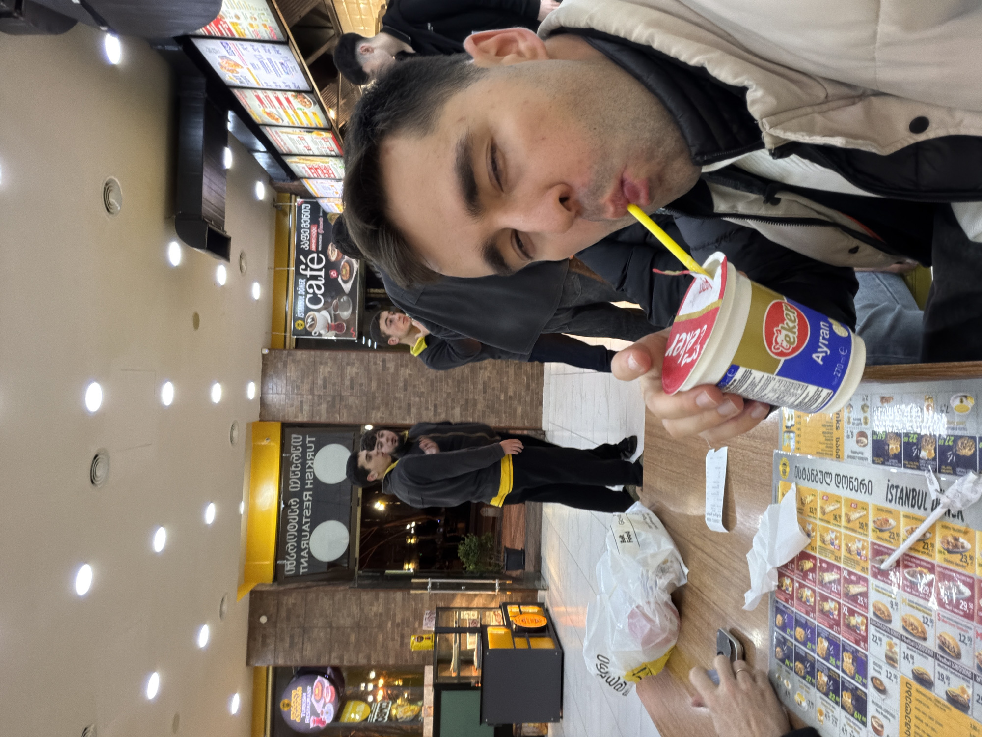 The image depicts the interior of a Turkish fast-food establishment in Tbilisi, Georgia, at night. In the foreground, a male with short dark hair, wearing a beige puffer jacket over a black one, looks directly at the camera while taking a selfie. He is actively drinking from a white plastic "Seker Ayran" cup with a yellow straw, his mouth pursed. His left hand holds the cup.
Seated at a light-colored wooden table, a partial view of another person's hands is visible near a silver smartphone. The table displays a laminated menu featuring "Istanbul Doner" and other food items with Georgian text and numerical prices, a crumpled white paper napkin, and a small white receipt. A clear plastic bag, with "Bolt Food" visible, containing unspecified items, rests on the table.
In the midground, to the left, two young males stand near the restaurant's glass entrance. Both wear dark jackets, one with a yellow undershirt, and appear to be observing the exterior, which shows a dark street scene with streetlights and tree silhouettes. Further to the right in the background, another male in a dark jacket is partially visible near the counter area.
The restaurant's interior features a light-colored tiled floor, brown brick-patterned walls, and a cream-colored ceiling with multiple recessed circular lights and air vents. Signage on the walls includes "TURKISH RESTAURANT" (mirrored) and "Istanbul Doner Cafe" with accompanying Georgian script. A glass display case with yellow accents is positioned to the left near the entrance. Backlit menu boards displaying food options are visible above the counter area on the right.