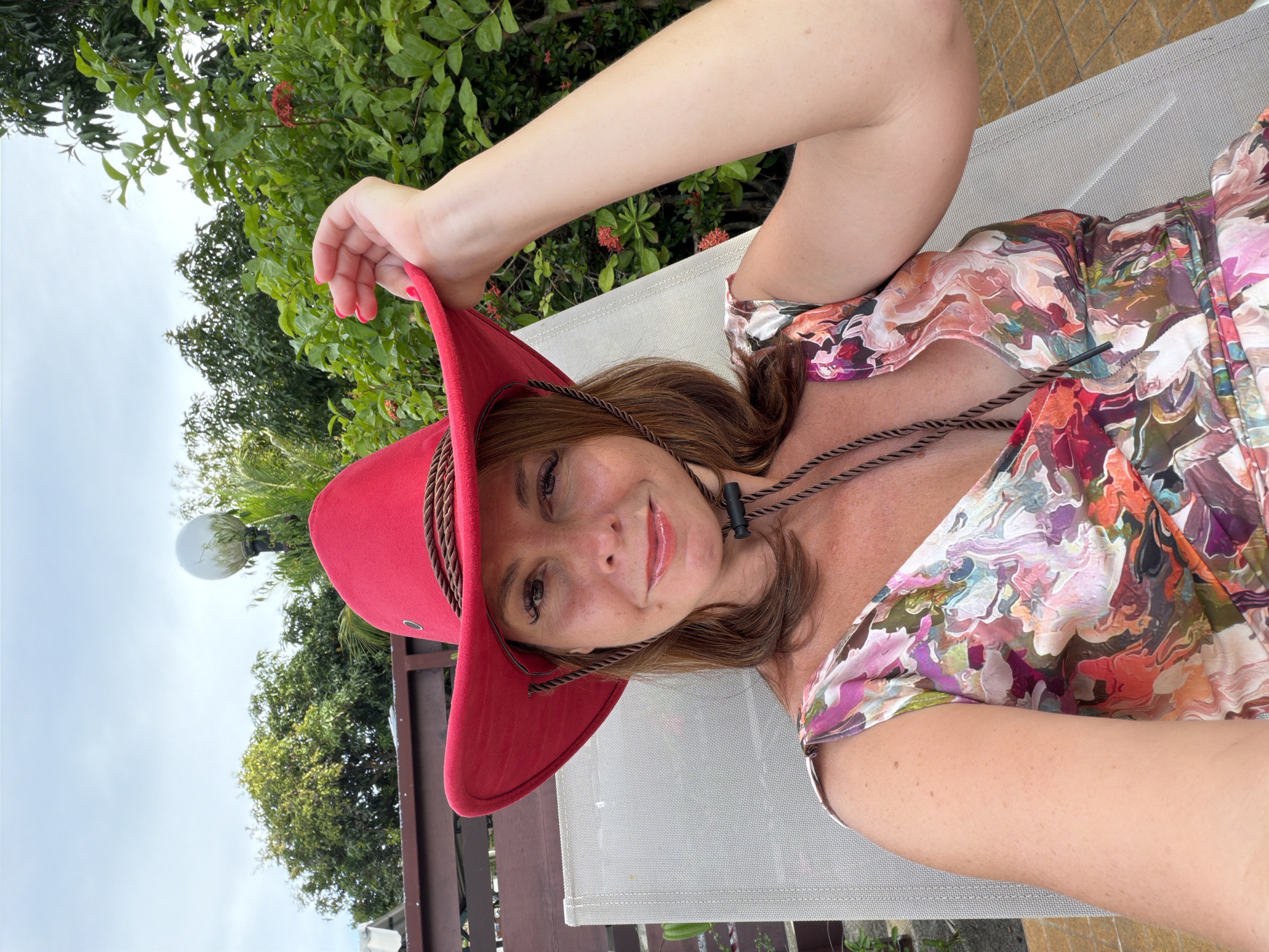 A woman wearing a red cowboy hat and a floral dress is sitting in a lounge chair outdoors. She is holding the brim of her hat with her right hand. The background shows lush green foliage and a cloudy sky. The location is identified as Baan Mae Nam, Thailand.