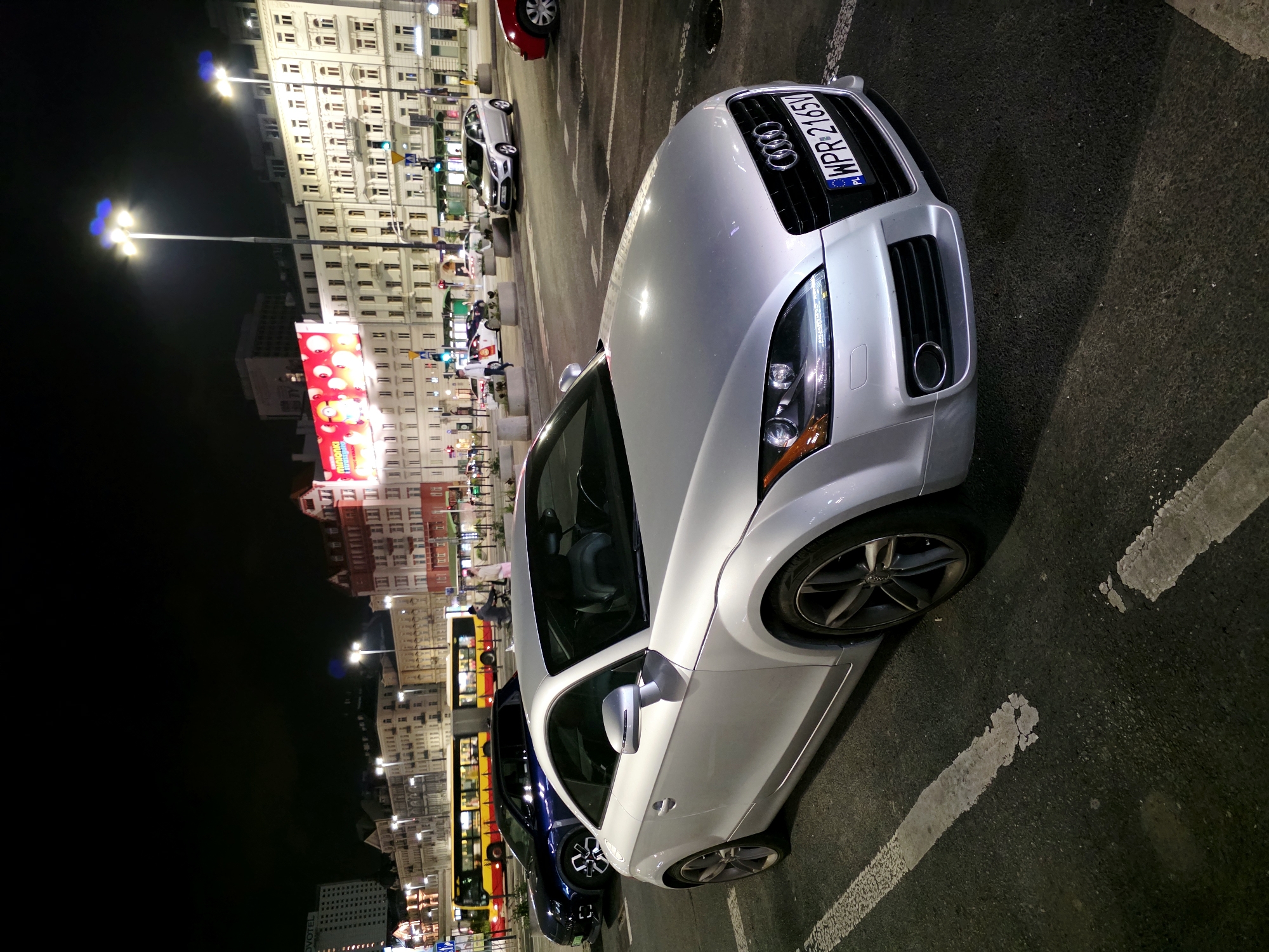 A silver Audi TT is parked in a lot at night on a city street. The car is in the foreground, angled to the right, showing its front end and passenger side. Behind it, a dark blue sedan is parked, and further back, a yellow and red bus is driving. The street is lined with buildings of various heights and architectural styles, some of which are illuminated. Streetlights are visible, casting a glow on the scene. The overall impression is a busy urban environment at night.