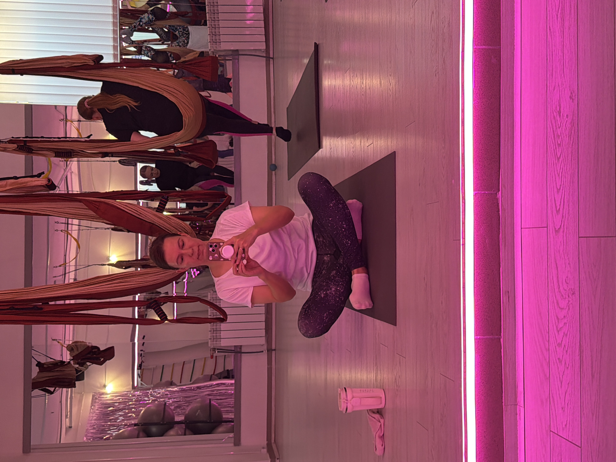 A woman in a white t-shirt and patterned leggings is sitting cross-legged on a yoga mat, taking a selfie with her phone. She is in a room with wooden floors and a pink neon light strip along the bottom of the wall. In the background, aerial yoga hammocks are hanging from the ceiling, and other people are visible in the reflection of a large mirror. The overall scene suggests a yoga or fitness studio. There is no information available to determine the location in Chaikovsky, Russia.