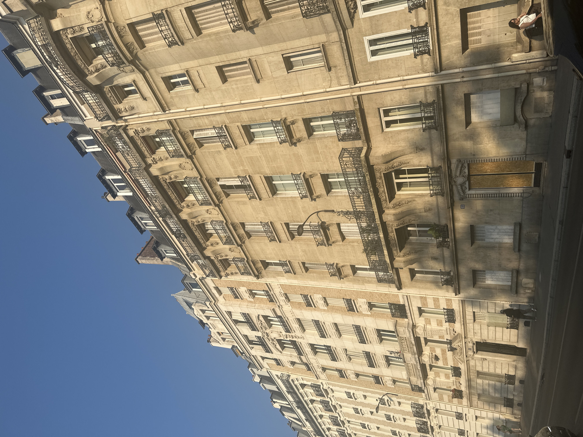 The image shows a street view of classic Parisian apartment buildings on a clear, sunny day. The buildings are tall, multi-story structures with ornate stone facades, mansard roofs, and numerous windows with wrought-iron balconies. The architecture is typical of Haussmannian-era Paris. The street is paved, and there is a sidewalk in front of the buildings.
On the sidewalk, there are a few people. Towards the left, a woman in a green dress is walking. In the middle of the sidewalk, a person in dark clothing is walking away from the camera. Towards the right, a woman in a white top and patterned skirt is sitting on a ledge, looking towards the right. There are also parked cars visible in the foreground. The sunlight casts long shadows, indicating it might be late afternoon or early morning.
The overall impression is a serene and picturesque urban scene in Paris, France.