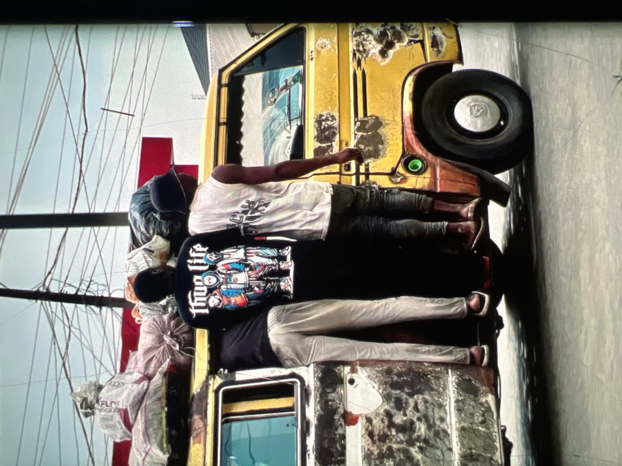 Three individuals are precariously positioned on the exterior of a yellow, rust-damaged commercial van in Lagos, Nigeria. Two men are partially visible inside the open doorway of the van, while a third man stands on the exterior, to the left, with his legs inside the van and his torso extending outwards. The van is laden with several large sacks and bundles of cargo secured to its roof. The individuals' clothing is casual, with one wearing a black t-shirt with a "Thug Life" graphic featuring cartoonish masked figures. The environment suggests a busy urban setting with overhead power lines and some buildings visible in the background. The van appears to be in motion or stopped in a street.