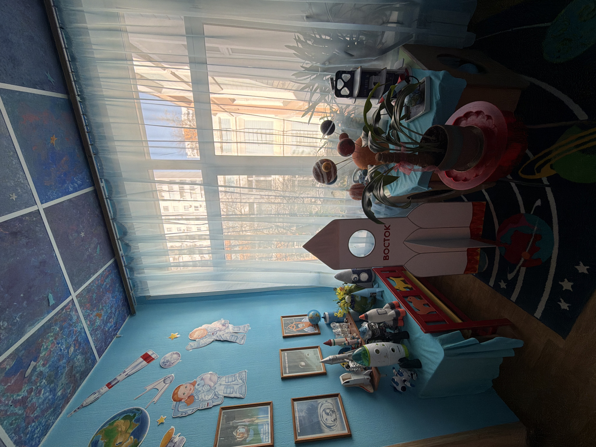 The image depicts a child's room or classroom decorated with space-themed elements. A wall is painted light blue and adorned with cutouts of astronauts, planets, and rockets. There are framed pictures depicting space-related subjects on the wall. A table is covered with a light blue cloth, upon which sit toy rockets, a globe, and decorative space items. A large model of the Vostok rocket is also present. A window behind the table is covered with sheer white curtains. Through the window, buildings and trees are visible. The room's location is likely in Khimki, Russia, judging from the name "BOCTOK" (Vostok) on the rocket model, written in Cyrillic script. The carpet in the room is dark blue with designs of planets and stars.