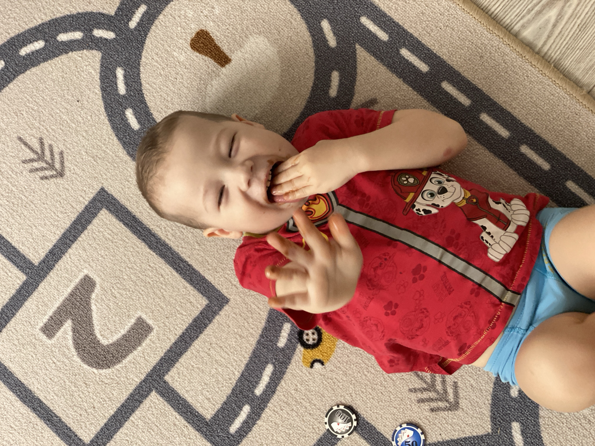 A young, light-skinned child with short, light brown hair is depicted lying on their back on a light-colored rug with a road map design. The scene takes place indoors in a location within the city of Sochi, Russia. The child is wearing a red short-sleeved t-shirt featuring a "Paw Patrol" character (Marshall, the Dalmatian firefighter) and blue shorts. Their eyes are closed, and their mouth is open in a wide expression, revealing teeth. The child's left hand is positioned inside their mouth, with fingers visibly coated in a brown substance. The right hand is raised and extended forward, palm facing the viewer, with all fingers splayed and also covered in the same brown substance. To the left of the child, on the rug, two poker chips are visible: one black chip marked with "100" and playing card motifs, and one blue chip marked with "50" and playing card motifs. The rug features dark grey road patterns with white dashed lines, a brown "2" numeral, brown arrow symbols, and a partial yellow vehicle graphic. The lower right portion of the image shows a transition from the rug to a light-colored wooden floor.