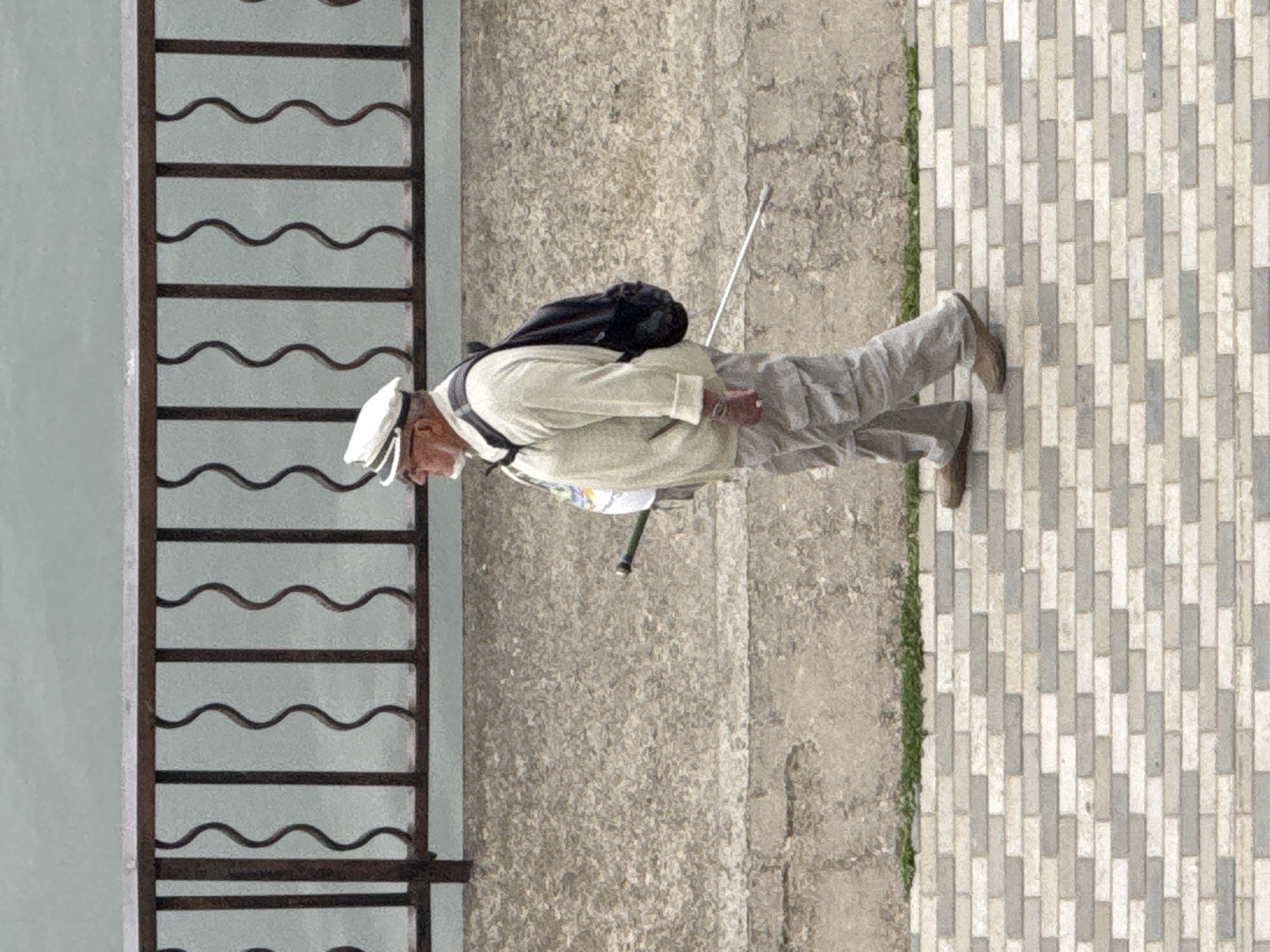 A Caucasian male walks in profile from left to right on a paved walkway. He is wearing a white peaked cap, eyeglasses, a light-colored jacket over a white shirt, and gray cargo pants. He has a black backpack on his back and is holding a walking stick in his right hand. The man is walking along a wall with a metal railing on top. The walkway is made of light-colored bricks. The background is a gray wall and a railing with wavy designs. The image has a slightly grainy texture. The scene is likely on the embankment of the Votka River in Votkinsk, Russia, given the context of the river and promenade-like setting.