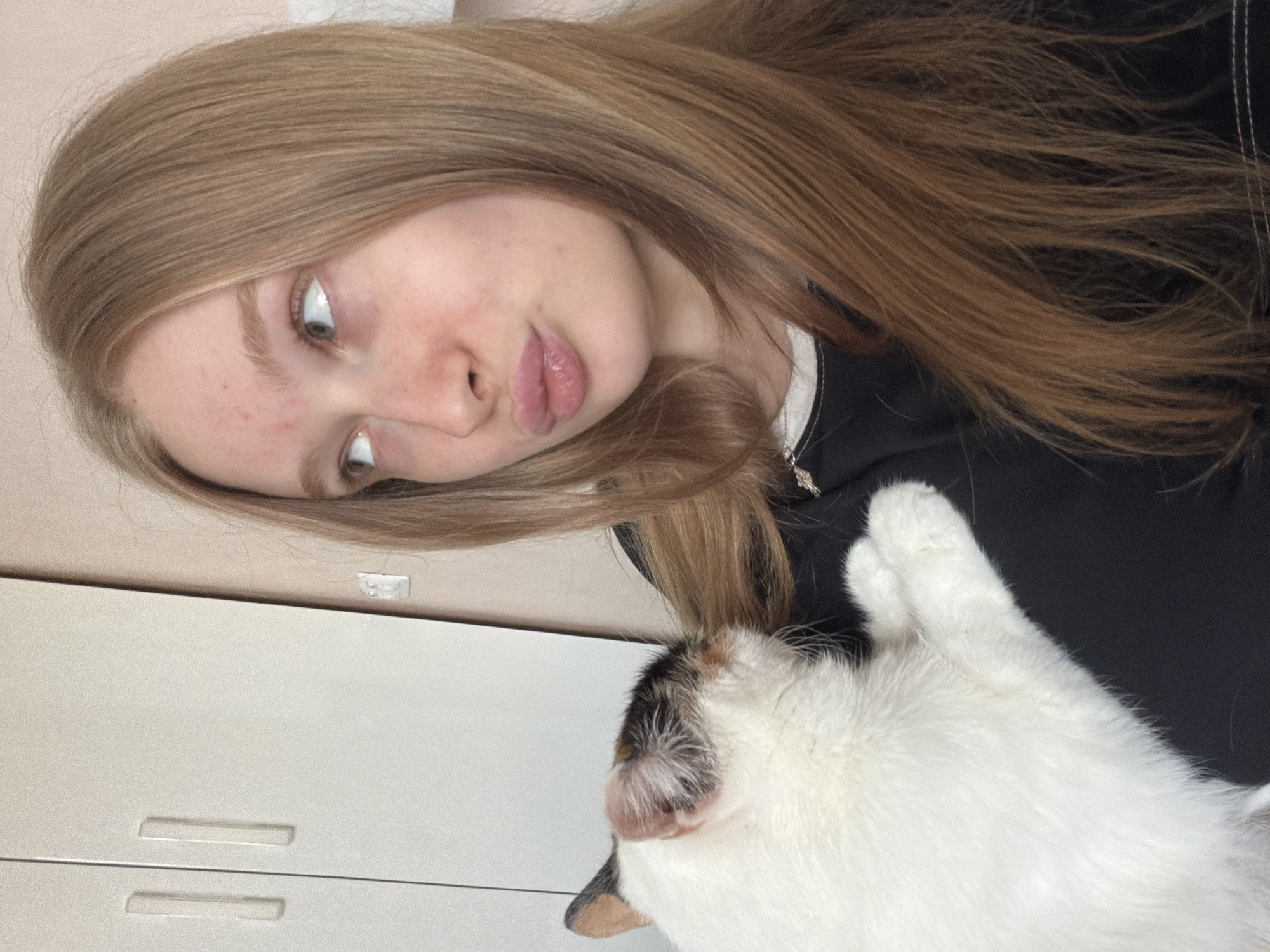 A woman with long, light brown hair is in the frame with a cat. The woman has fair skin and light eyes, and she is wearing a black top with a white neckline. She is looking down at the cat with a slight pout. The cat is mostly white with patches of black and brown on its head. The background includes a white cabinet with horizontal grooves. The location is Rossiyskiy, Russia.