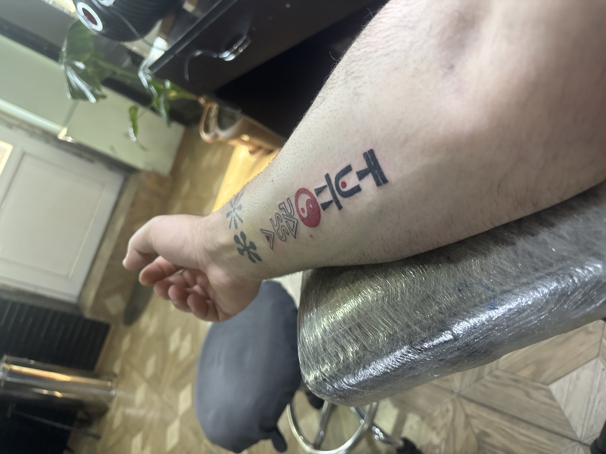 A medium shot depicts a person's forearm resting on a chair. The forearm has several tattoos. The most prominent tattoo is a sequence of symbols, including what appears to be a stylized Yin and Yang symbol colored red. Above it is a symbol similar to a "U" with a vertical line bisecting it, and to the left are three other symbols including a flower-like symbol. The arm is positioned over a stool covered in clear plastic wrap.
The background shows elements of an indoor environment. A kitchen-style floor and a kitchen with a gray stool are visible. The location appears to be in Tbilisi, Georgia.