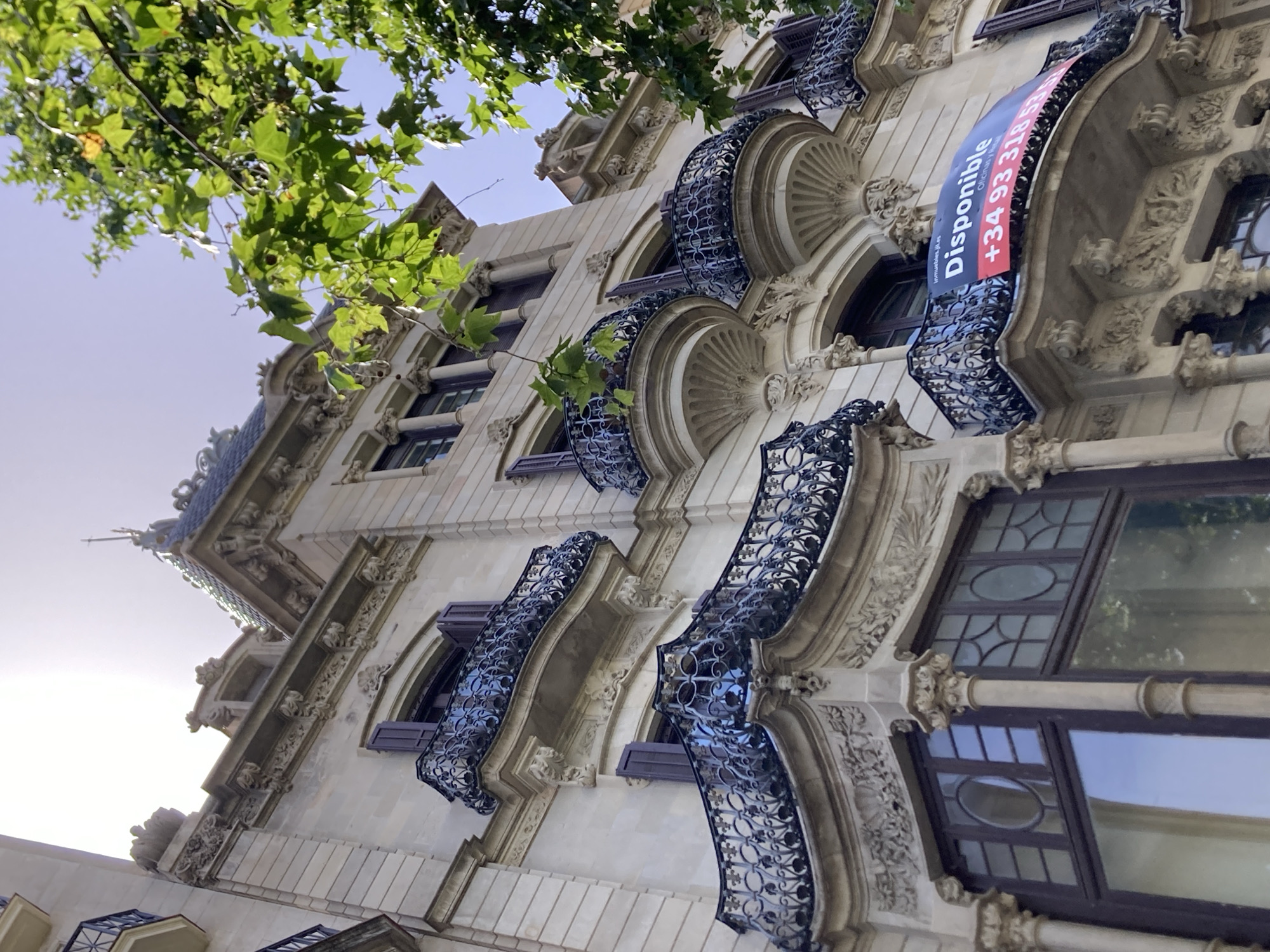 A low-angle shot captures the ornate facade of a building in Barcelona, Spain, under a bright sky. The architecture features detailed stonework, including decorative carvings around windows and balconies. Several wrought-iron balconies with intricate scrollwork jut out from the building. A banner advertising "Disponible" (Available) with a phone number is draped across one of the balconies. Green leaves from an overhanging tree frame the top right portion of the image. The building appears to be residential or office space.