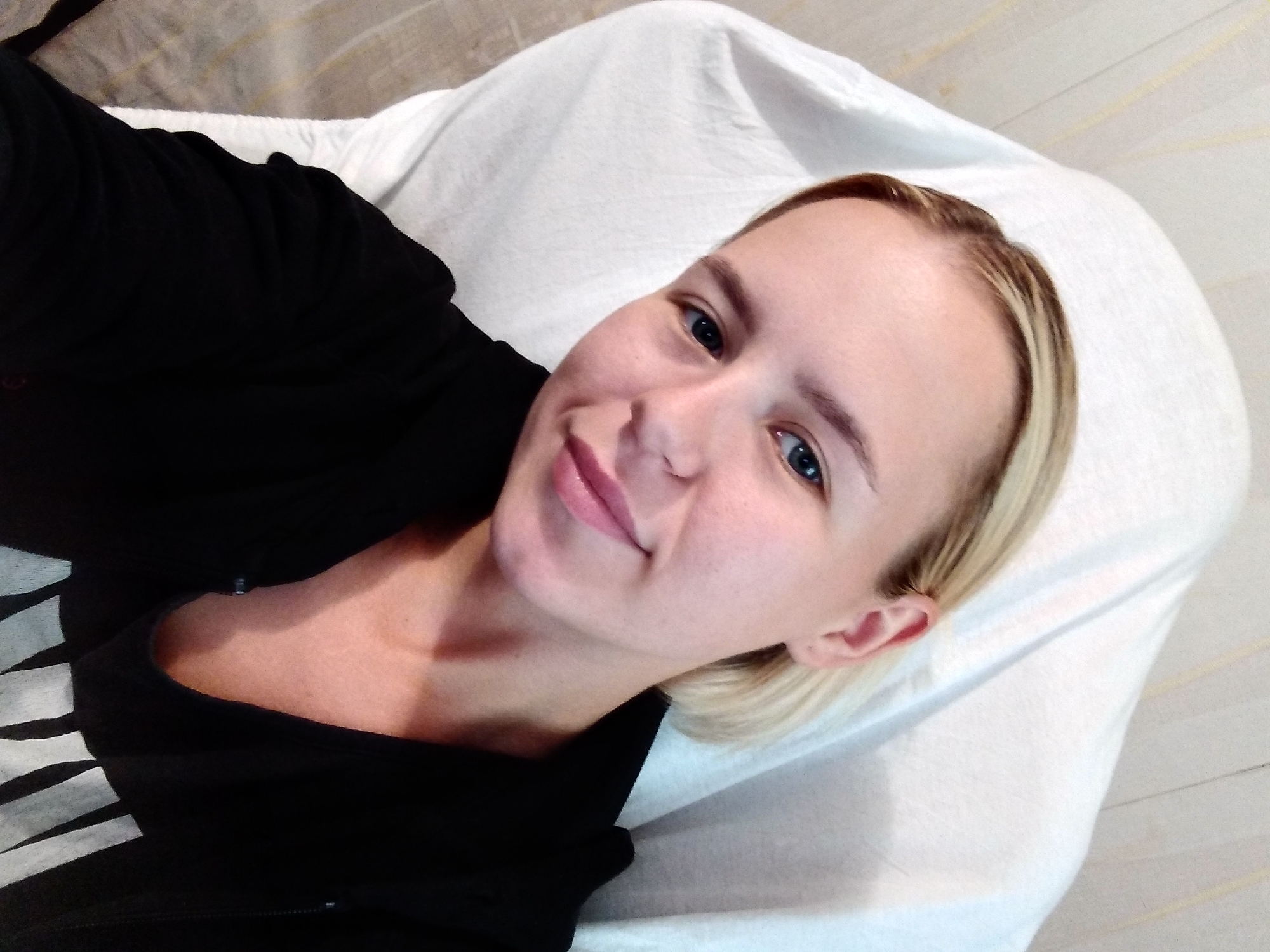 A woman with blonde hair and blue eyes is lying down on a white surface, possibly a bed or a treatment table. She is wearing a black top. Her head is tilted to the side, and she is looking at the camera with a slight smile. The lighting suggests it is daytime. The background is a neutral-colored wall with some textured lines. The overall impression is a casual and relaxed portrait.