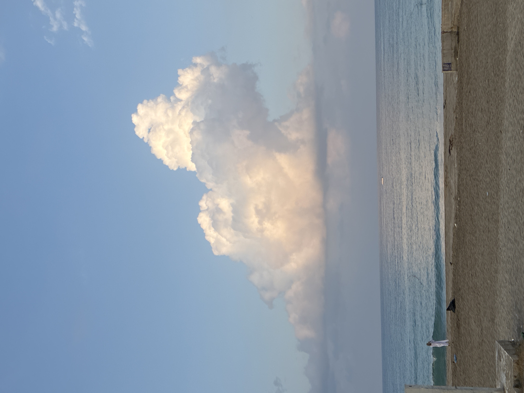 A wide shot captures a serene beach scene along the coast of Alanya, Turkey. The sky is a clear, pale blue with a few wispy clouds scattered near the horizon, dominated by a large, towering cumulus cloud formation. The sea is a tranquil turquoise, with gentle waves rolling onto the sandy shore.
On the beach, a solitary figure dressed in white stands near the water's edge, appearing to be taking a photograph. Further down the beach, a dark object, possibly a piece of luggage or a shadow, rests on the sand, and a small bird stands nearby. In the foreground, the sandy beach is relatively clear, with some concrete structures and debris visible at the edges. A small white boat or marker is visible on the water in the distance. The overall atmosphere is calm and peaceful, typical of a late afternoon or early morning on the Mediterranean coast.