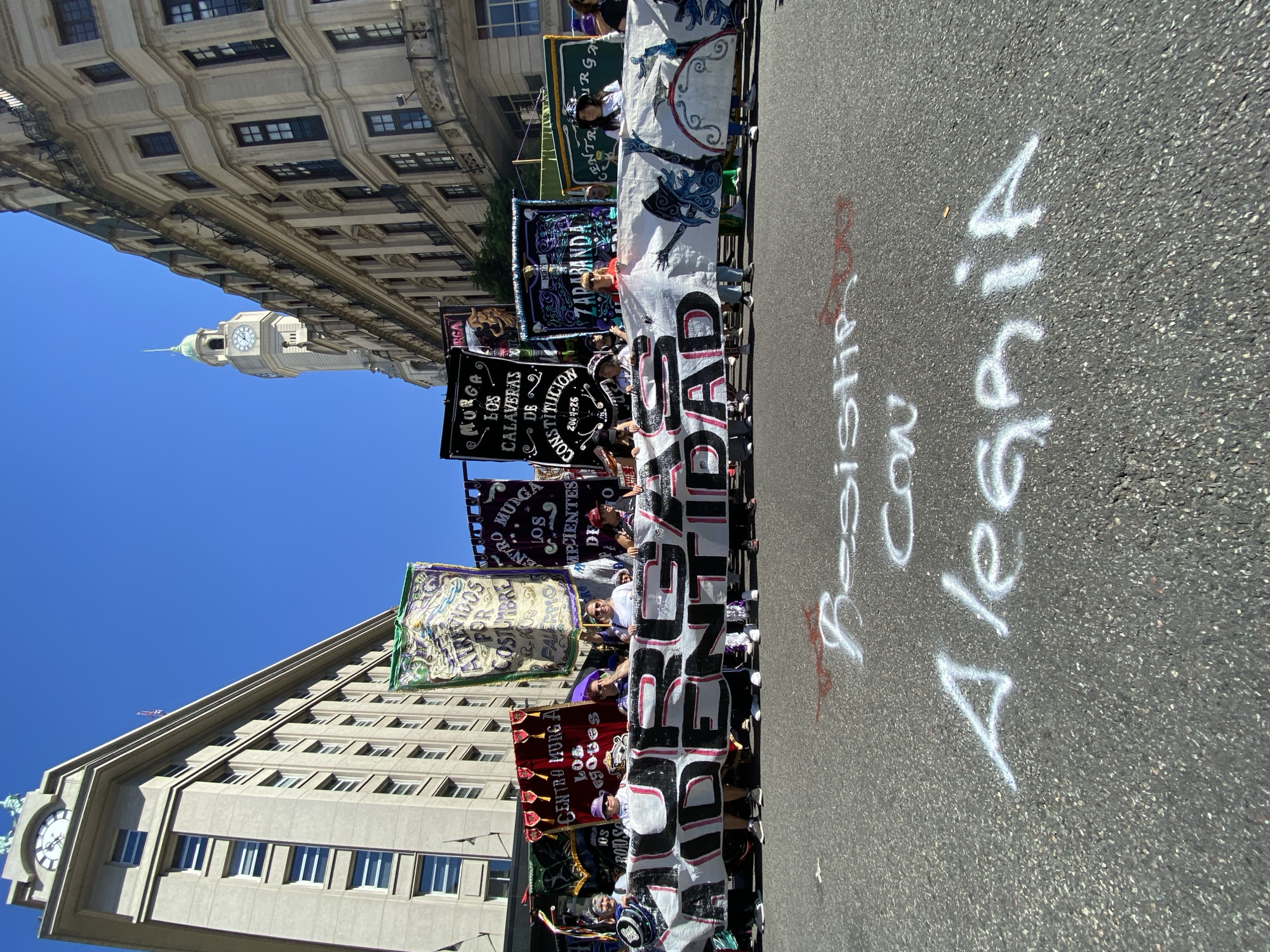 This image captures an outdoor street scene in the Montserrat neighborhood of Buenos Aires, Argentina, specifically along Diagonal Julio A. Roca (Diagonal Sur) or Hipólito Yrigoyen street.
Approximately 15-20 adults are positioned across an asphalt road in the middle ground, holding a large white fabric banner with black and red text, prominently displaying "SOLIDARIDAD" and partially "MURGA ARGENTINA." Behind this, numerous elaborate, colorful flags and banners, characteristic of "Murga" groups, are held upright, identifying organizations such as "CENTRO MURGA LOS NEGOBES," "MURGA LOS CALAVERAS DE CONSTITUCION," "ATREVIDOS POR COSTUMBRE PALERMO," and "ZARABANDA." The individuals are casually dressed, with some wearing hats.
In the foreground, white spray paint on the asphalt spells "Resistir Con Alegría" (Resist With Joy), with additional red spray paint visible. The scene is framed by multi-story buildings under a clear blue sky with bright sunlight. On the left, a large, light-colored building with a prominent clock tower at its peak is identifiable as the Palacio de la Legislatura de la Ciudad de Buenos Aires. On the right, an ornate, classical-style building with balconies and dark-framed windows lines the street. In the distance, above the right building, a slender clock tower with a green domed spire is visible, likely the Cabildo de Buenos Aires or another historical building in the vicinity of Plaza de Mayo. The street appears clear of vehicular traffic.