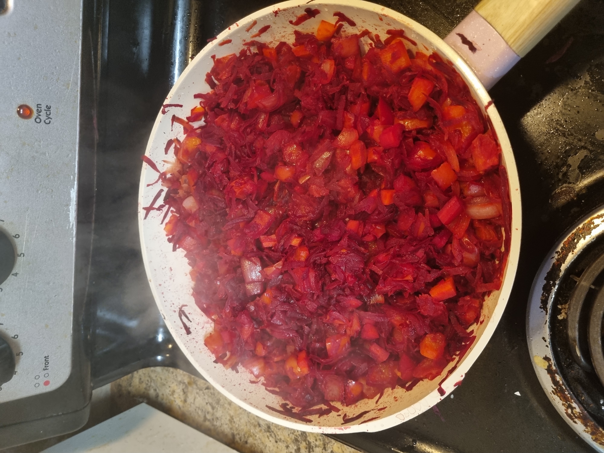 A close-up overhead shot shows a white and pink-handled skillet filled with a steaming mixture of shredded beets, diced red bell peppers, and onions, being cooked on a black electric stovetop. The stovetop has two burnt coil burners, one of which is partially visible in the lower left corner of the frame. The backsplash behind the stovetop is made of speckled granite. The stove's control panel is visible at the top, with markings for "Front" and "Oven Cycle" illuminated, indicating the stove is in use. The scene is domestic and appears to be a kitchen. The location is identified as Vancouver, United States of America.