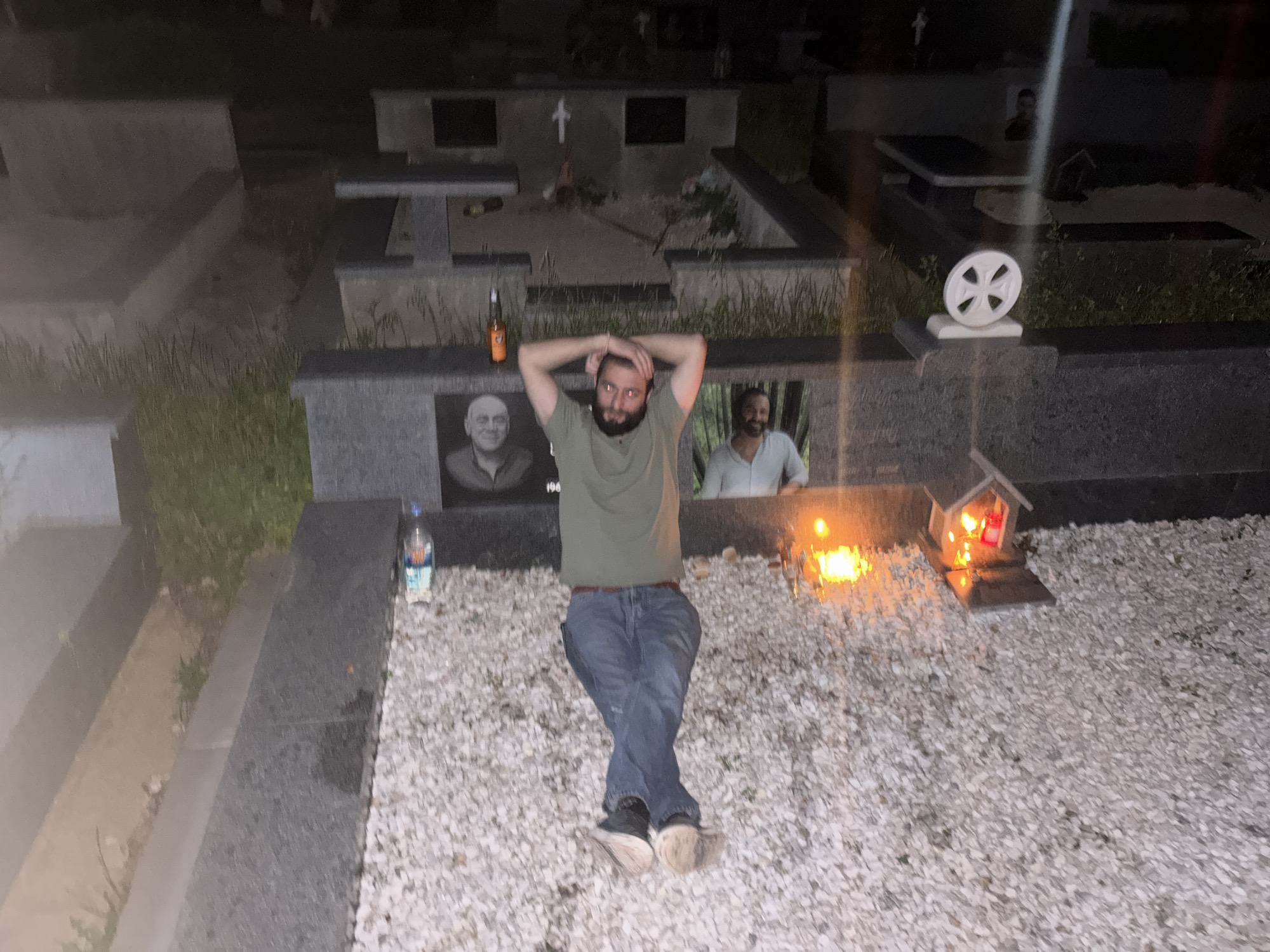 A man is lying down in a cemetery in Tbilisi, Georgia, with his hands behind his head and his legs crossed. He is wearing a green t-shirt, blue jeans, and black and white shoes. There are gravestones and a small memorial with candles in the background. A bottle of water and a beer bottle are also visible on the ground. The scene appears to be illuminated by artificial light.