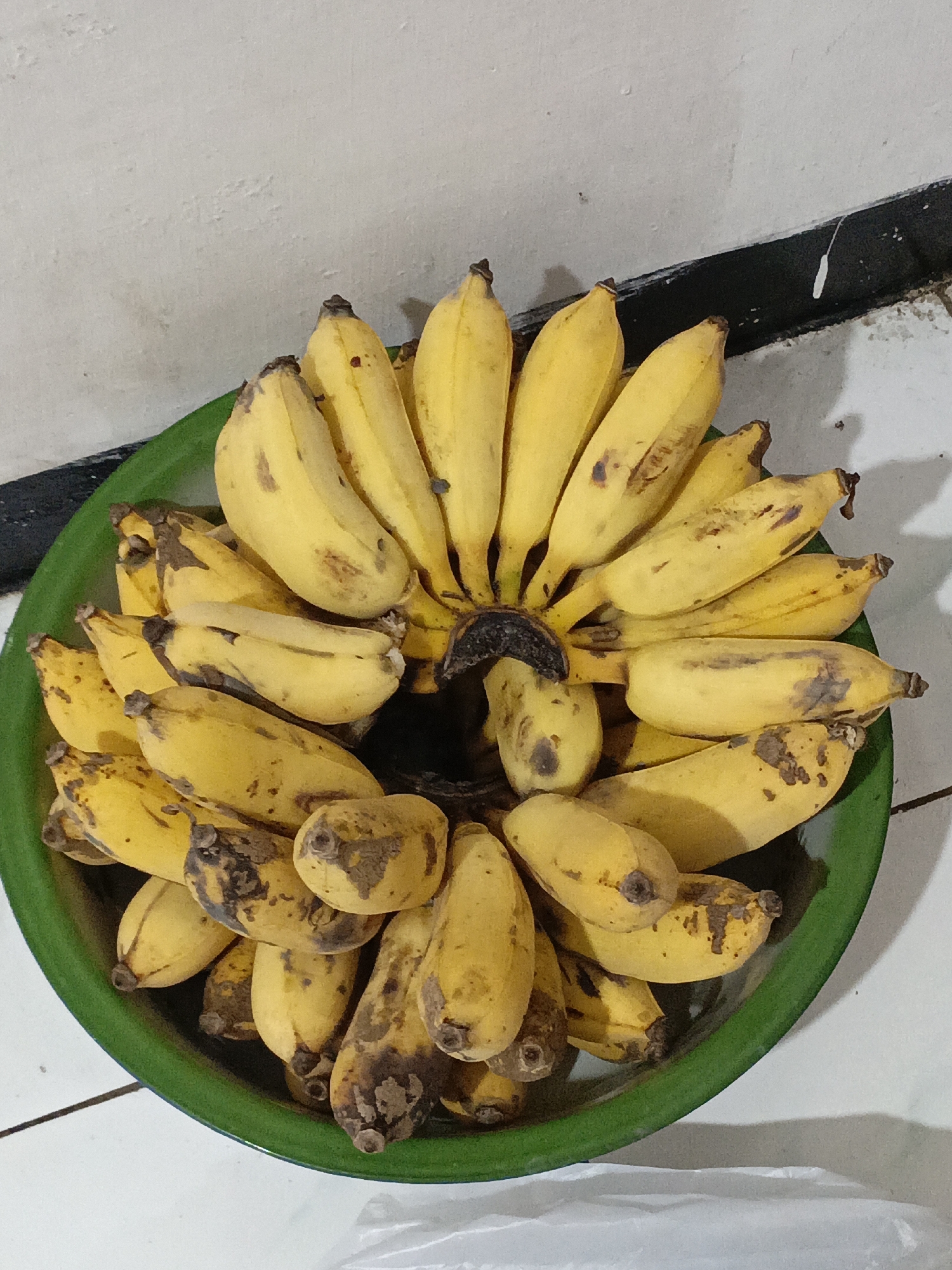 A bunch of ripe bananas is arranged in a green plastic bowl. The bananas are yellow with some brown spots, indicating ripeness. The bowl is placed on a white tiled surface, next to a wall that is partially painted white and partially has a black stripe near the floor. The scene appears to be indoors, possibly in a home kitchen or dining area in Salatiga, Indonesia. There are no people visible in the image, and the focus is solely on the fruit and its immediate surroundings. The lighting is somewhat dim, casting shadows on the wall and floor.