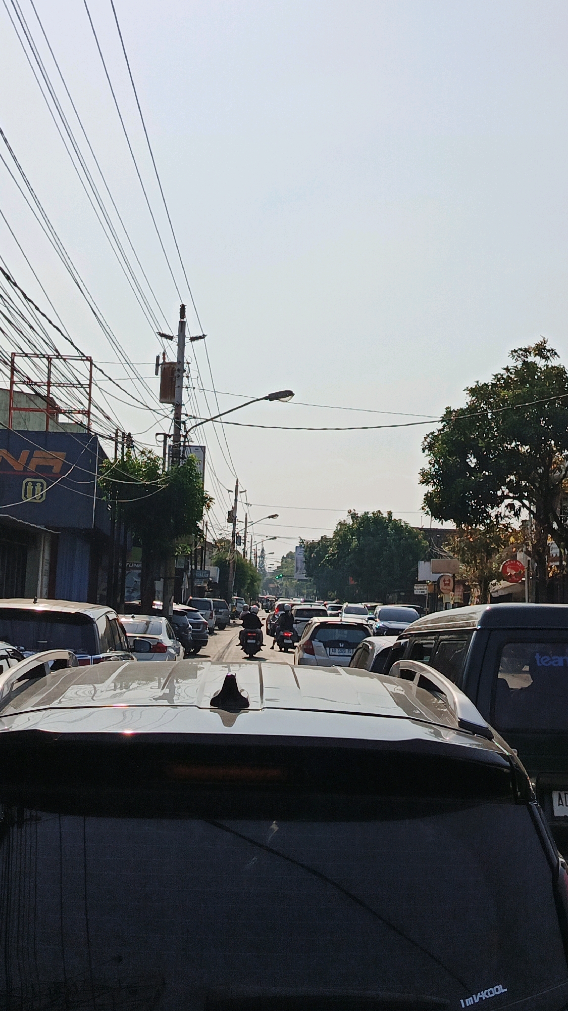 A traffic jam is depicted on a street in Surakarta, Indonesia, under a bright, hazy sky. The foreground is dominated by the rear of a dark-colored SUV, with its roof antenna and rear window visible. Behind it, other vehicles, including a silver SUV, are tightly packed on the road. A motorcycle is moving through the traffic. Buildings line the street, with one on the left featuring signage and utility poles with numerous electrical wires stretching across the frame. Trees are present on the right side of the street. The scene suggests a typical urban congestion.