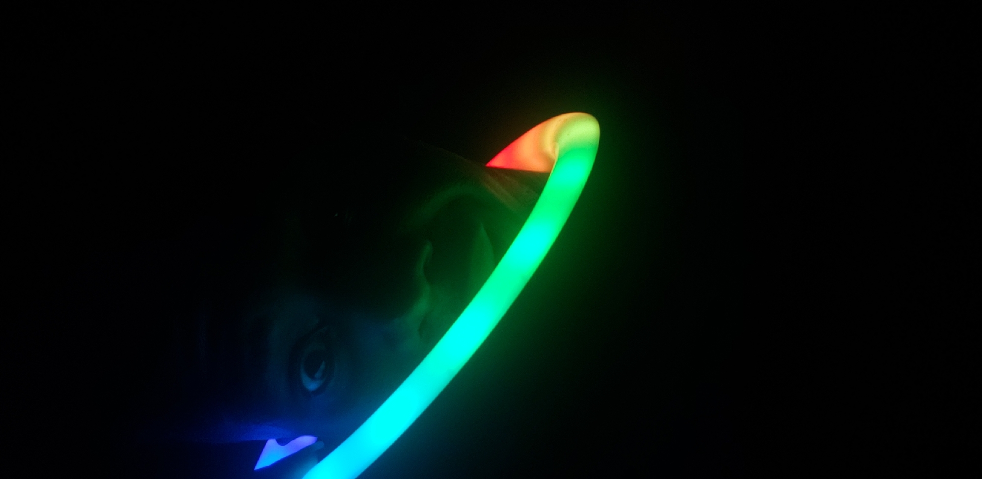 A close-up, low-angle shot captures a person's face in the dark, illuminated by a glowing, curved object that transitions from blue to green to red. The object, possibly a glow stick, is positioned in front of the person, obscuring part of their face and casting vibrant colored light. The person's eye is wide open, and their mouth is slightly agape, suggesting surprise or excitement. The background is entirely black, creating a stark contrast with the illuminated subject. The lighting suggests a dimly lit environment, possibly indoors or at night. There are no identifiable landmarks or features that would indicate the location in Narayi, Nigeria.