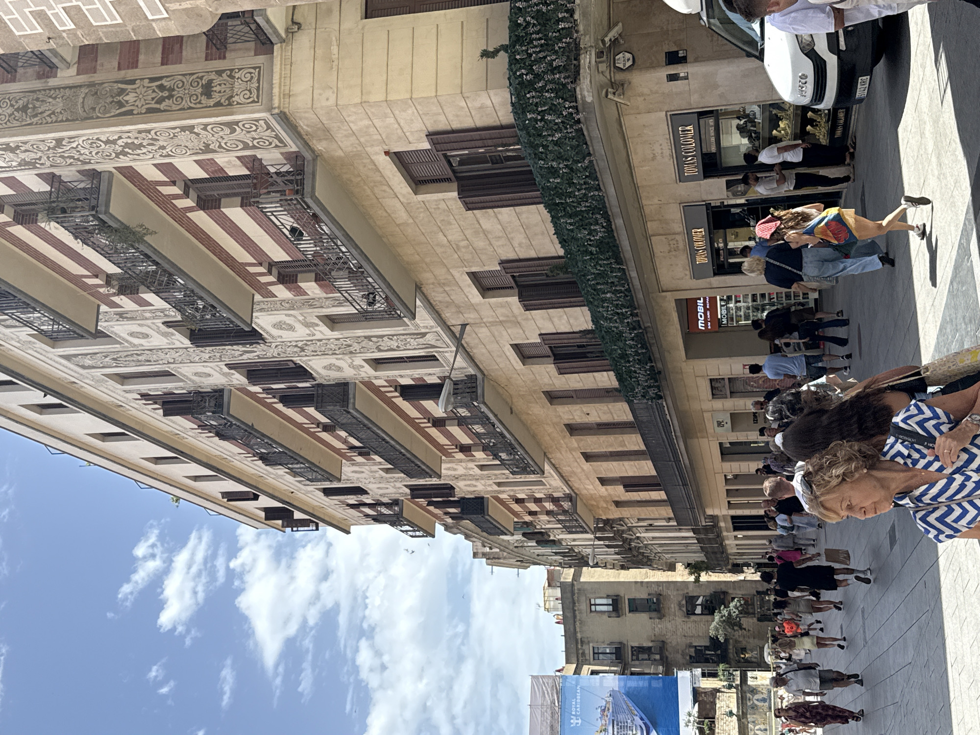 This media file captures a street scene in Barcelona, Spain, on a sunny day. A group of people are walking along a pedestrianized street lined with traditional European architecture. The buildings feature ornate facades with balconies and decorative elements.
Several shops are visible along the street, including "TOMAS COLOMER JEWELLERS SINCE 1876" and "MOBILE SIM CARD REPAR." A white car is parked on the side of the street, partially obscuring one of the shop fronts. People are dressed in summer attire, suggesting warm weather. The presence of a cruise ship advertisement for "ROYAL CARIBBEAN" hints at a tourist area. The overall mood appears to be one of casual strolling and shopping, typical of a vibrant city center. The image is oriented vertically, with the sky visible at the top and the street stretching downwards.