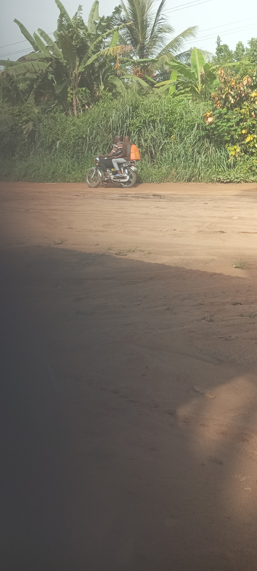 The image depicts a motorcycle with two people riding on a dirt road in Amauzu, Nigeria. The background consists of lush green vegetation, including banana trees and tall grass. The road is sandy and uneven. The foreground is out of focus, obscuring details.