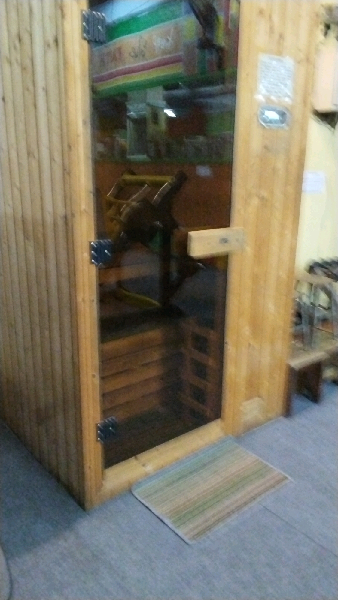 The image shows a wooden sauna room with a glass door. Inside the sauna, there are wooden benches and some equipment that is difficult to discern. A striped rug is placed on the floor in front of the sauna door. The background is blurred, but appears to be a commercial establishment with signage. Based on the provided crops, which show what appears to be a sign with Arabic script in the background, and the specific mention of the city of Mojokerto, Indonesia, this sauna is likely located in a public establishment such as a gym, spa, or recreational facility within Mojokerto. There are no people visible in the image.